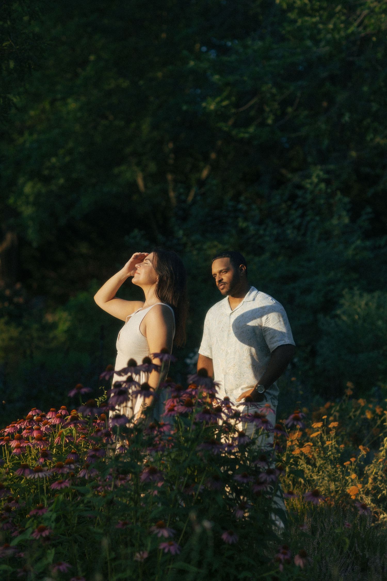 Couple standing among pink wildflowers in golden hour light, surrounded by lush green trees in a romantic outdoor setting.