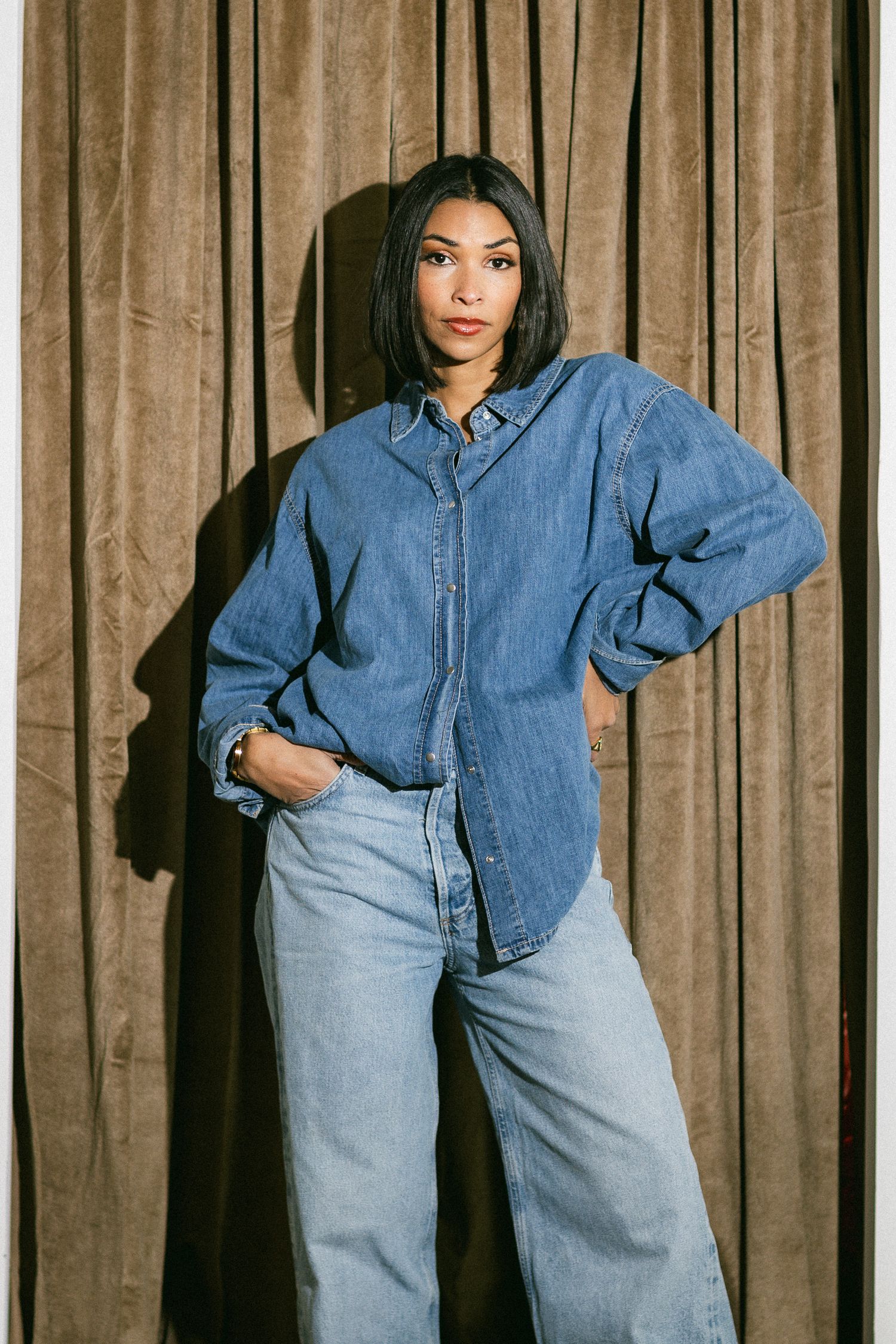 Woman in oversized blue denim shirt and wide-leg jeans posing before brown curtain backdrop.