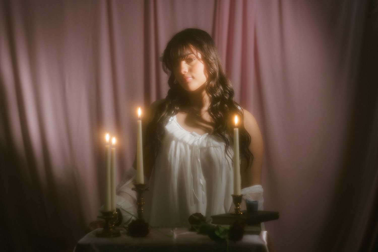Woman in white dress sitting before lit candles with pink curtain backdrop.