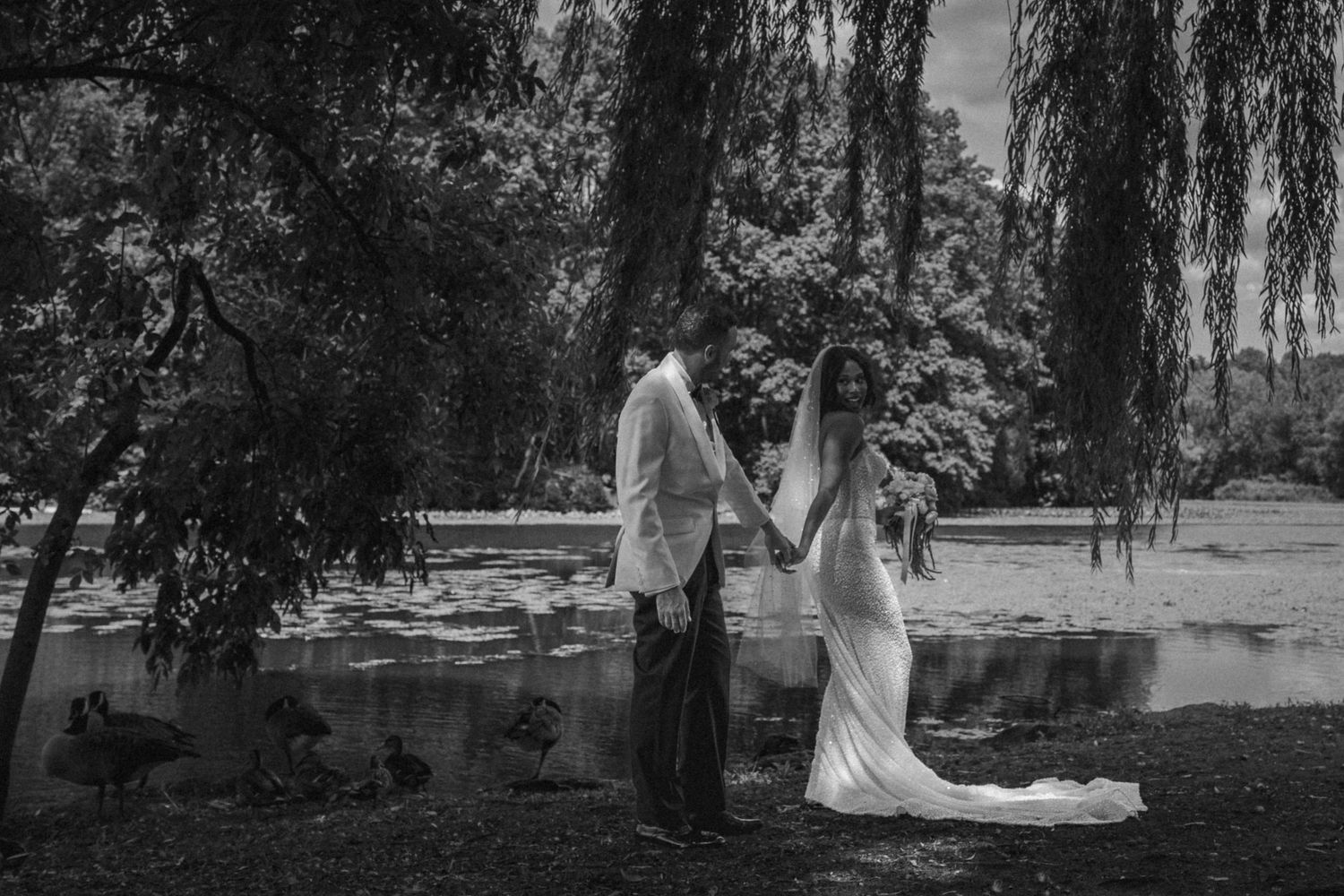 Black and white photo of a bride and groom holding hands near a willow tree by a lake with ducks.