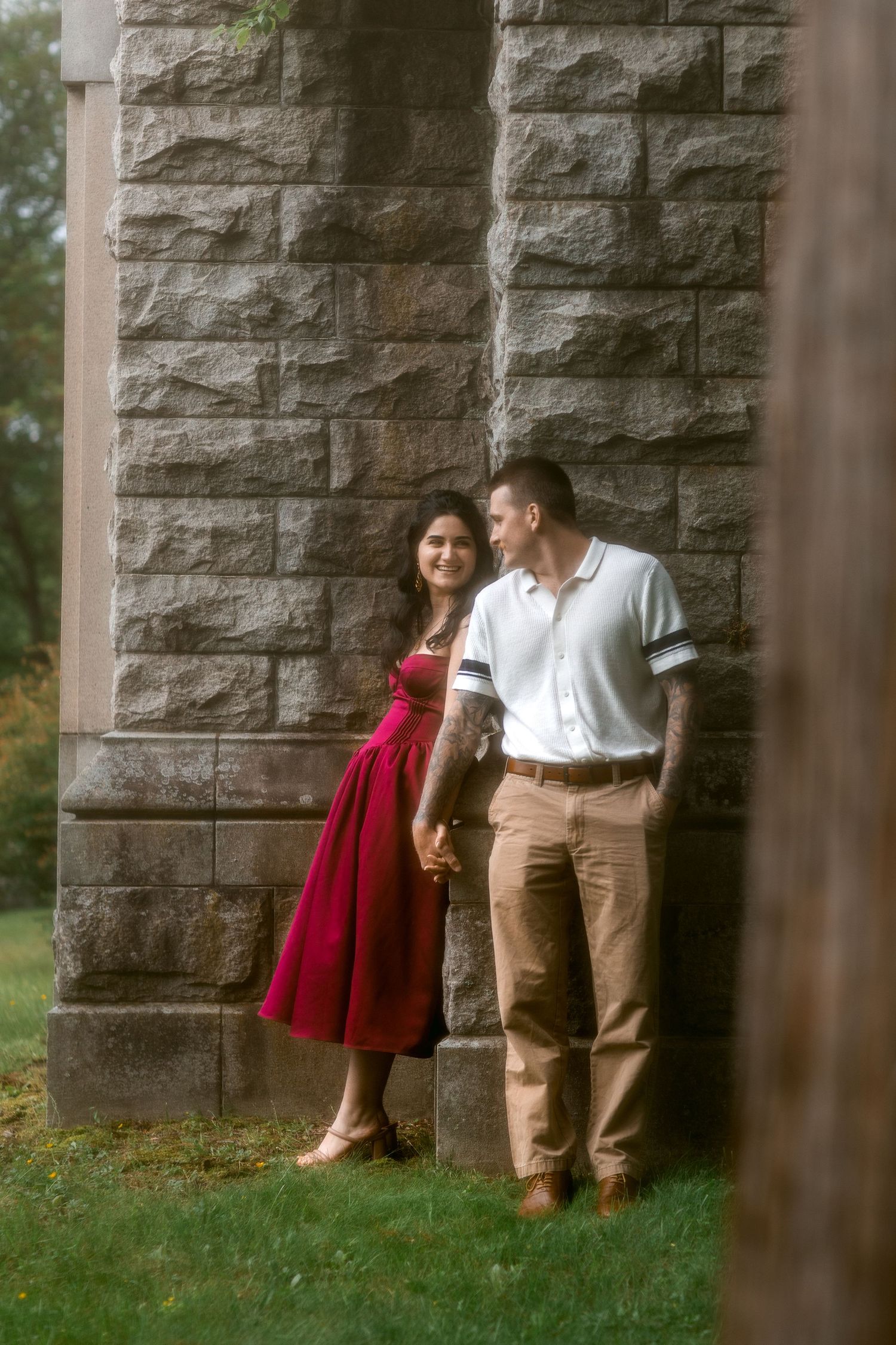 Couple holding hands by stone pillars, woman in red dress, man in white polo and khaki pants, outdoor setting.