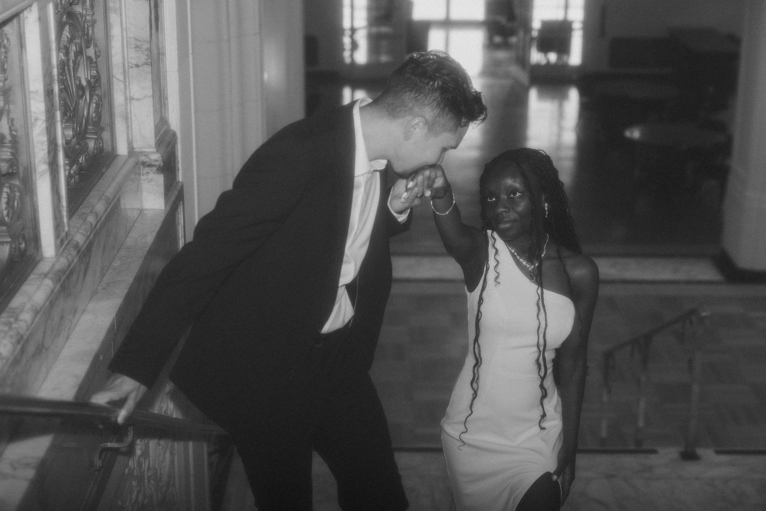 Black and white photo of couple sharing tender moment on staircase, man in suit kissing womans hand.