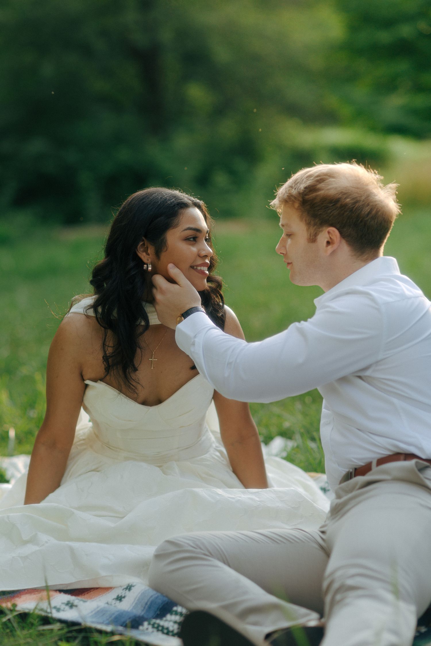 Bride and groom sitting on blanket in green park, groom tenderly touching bride's face as she smiles.