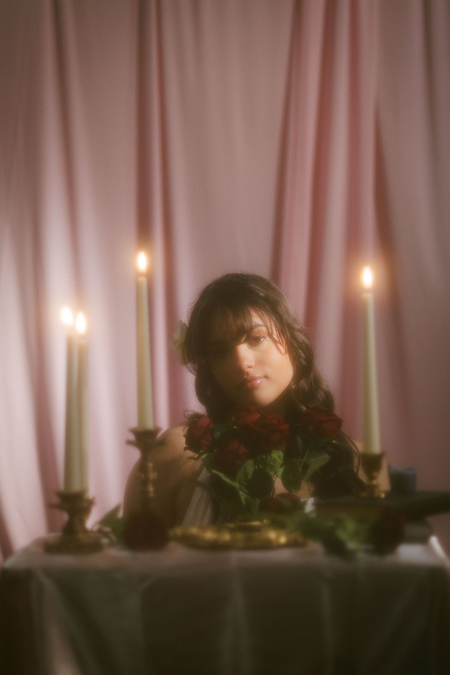 Dark-haired woman leaning over candlelit table with red roses against soft pink draped backdrop.