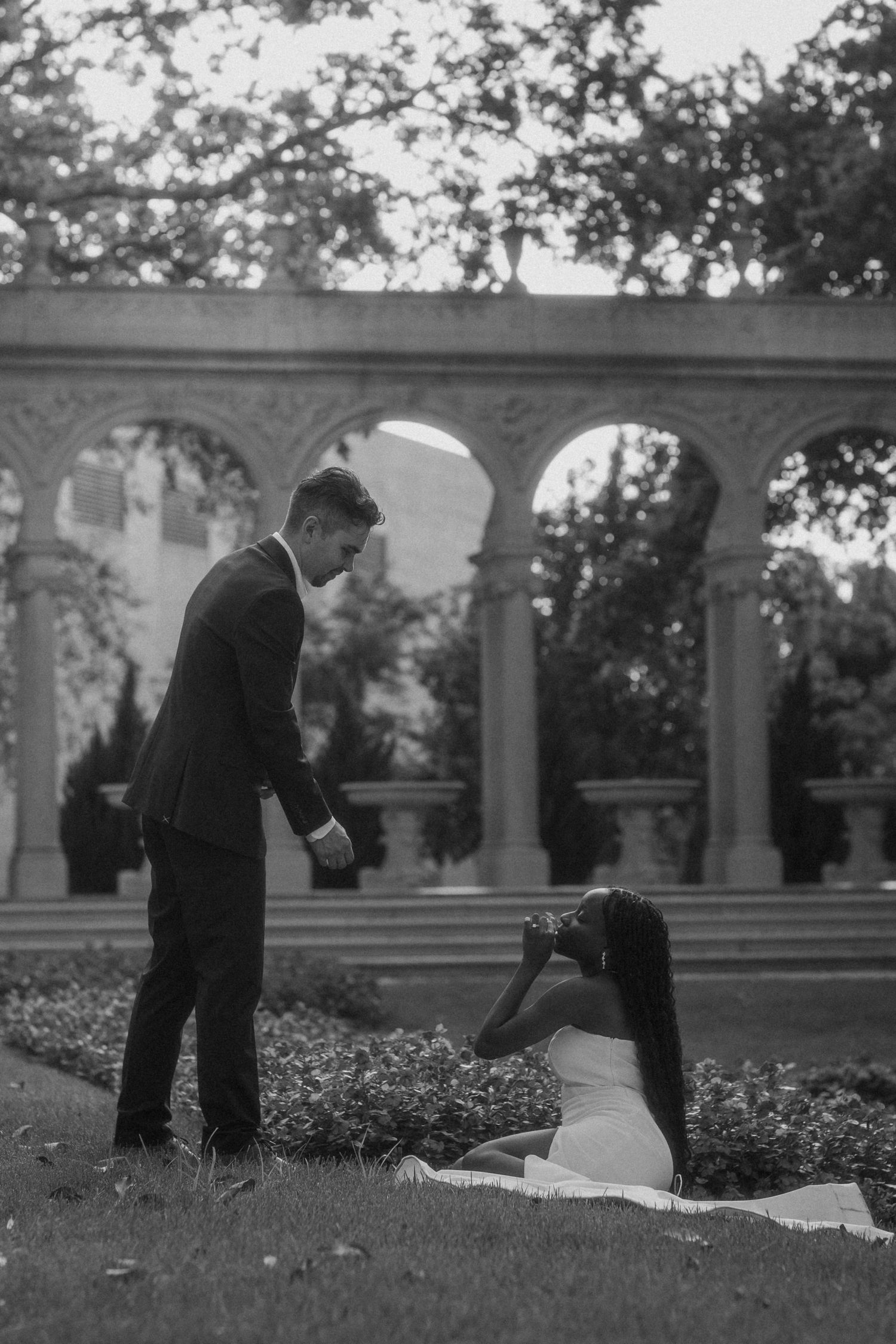 Black and white outdoor wedding photo of groom standing over seated bride in elegant garden with stone arches backdrop.