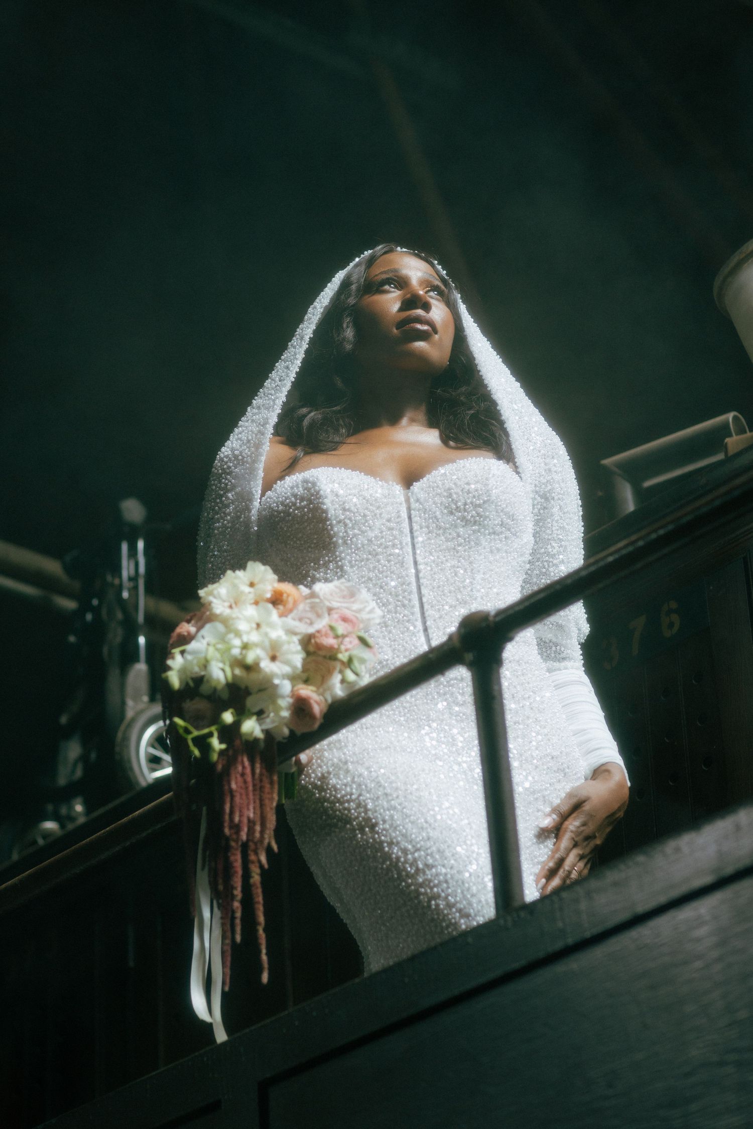 Stunning bride in white long-sleeve sequined gown and veil holds bouquet while standing on staircase.