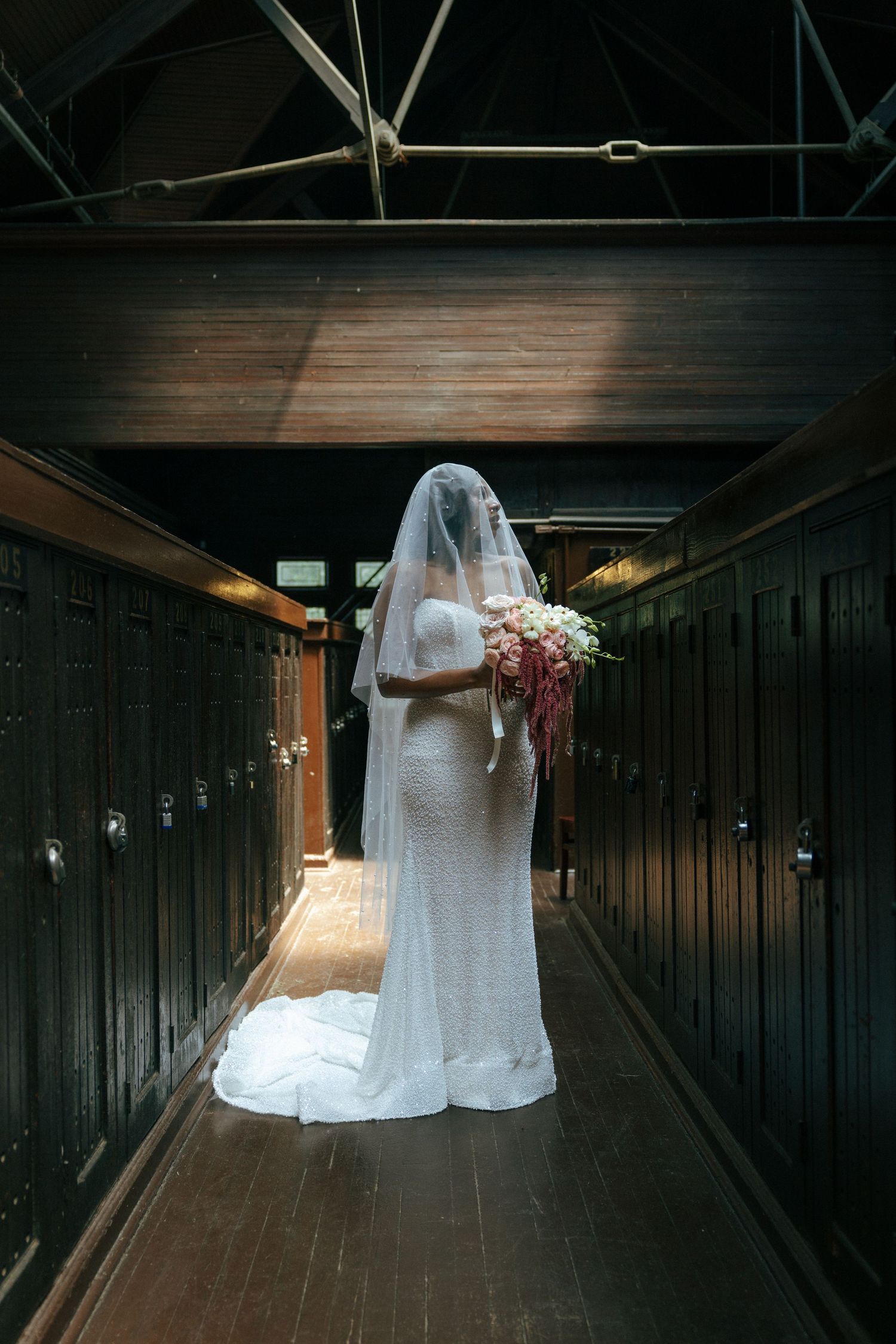Bride in white gown and veil holding bouquet in dark locker room hallway.