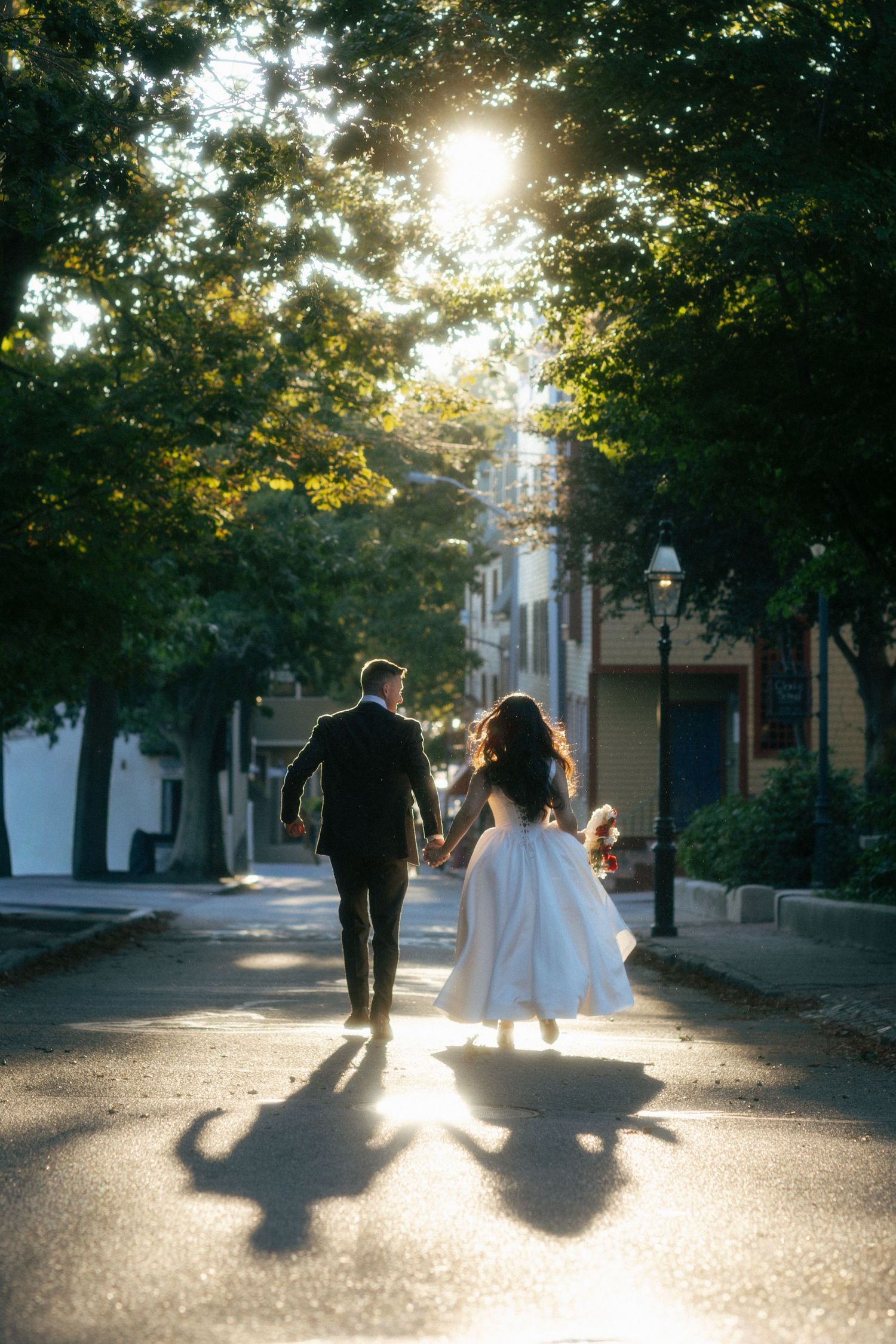 Bride and groom walk hand-in-hand down sunlit tree-lined street, casting long shadows on pavement.