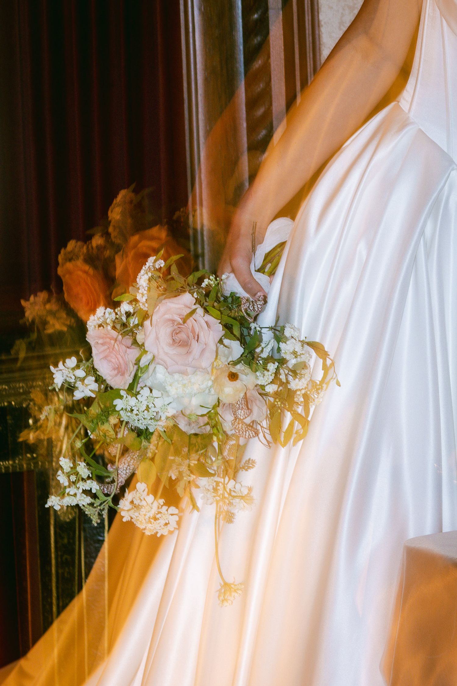 Bride in white gown holding a romantic bouquet of blush pink roses and white wildflowers near a window.