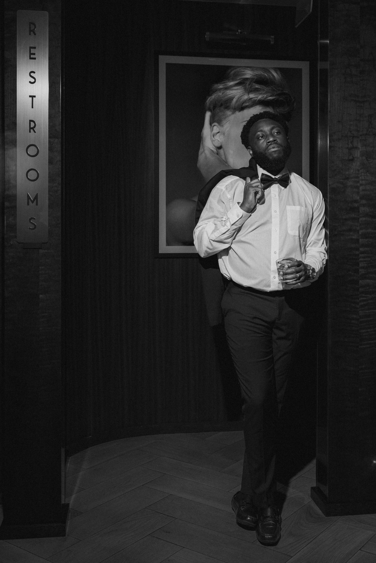 Elegant man in white shirt and bow tie poses near restrooms sign in black and white portrait.
