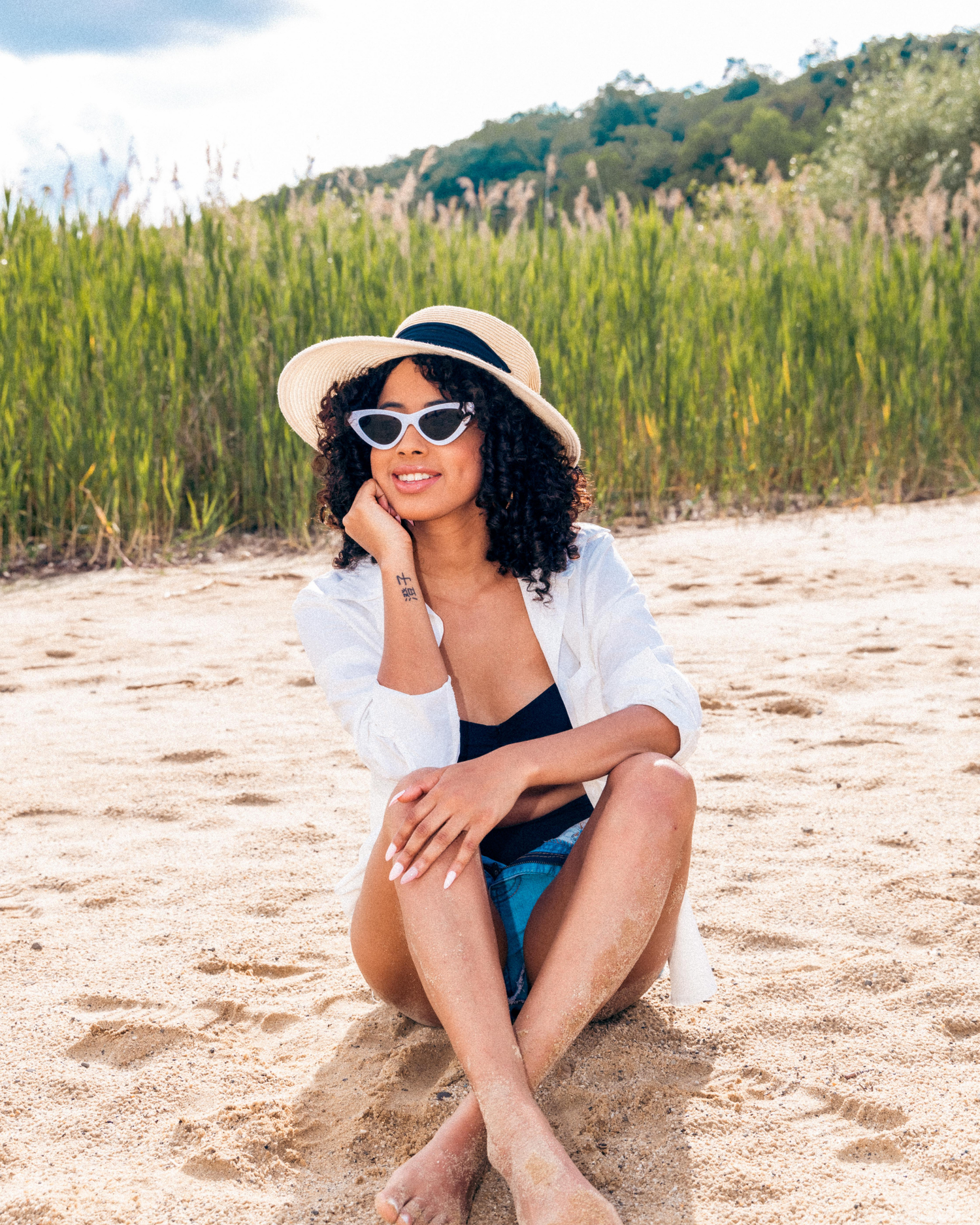 Young woman in straw hat and white sunglasses sitting on sandy beach, wearing white shirt and black swimsuit.
