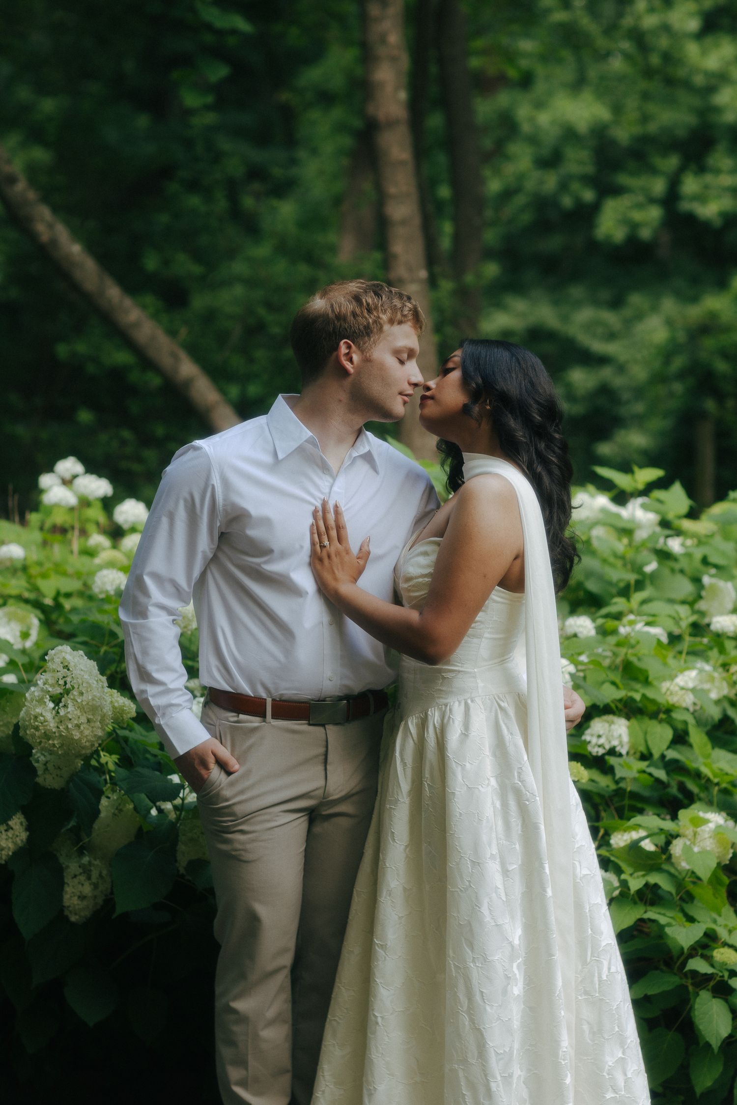 A couple shares a romantic kiss among white hydrangeas in a lush green garden, dressed in wedding attire.