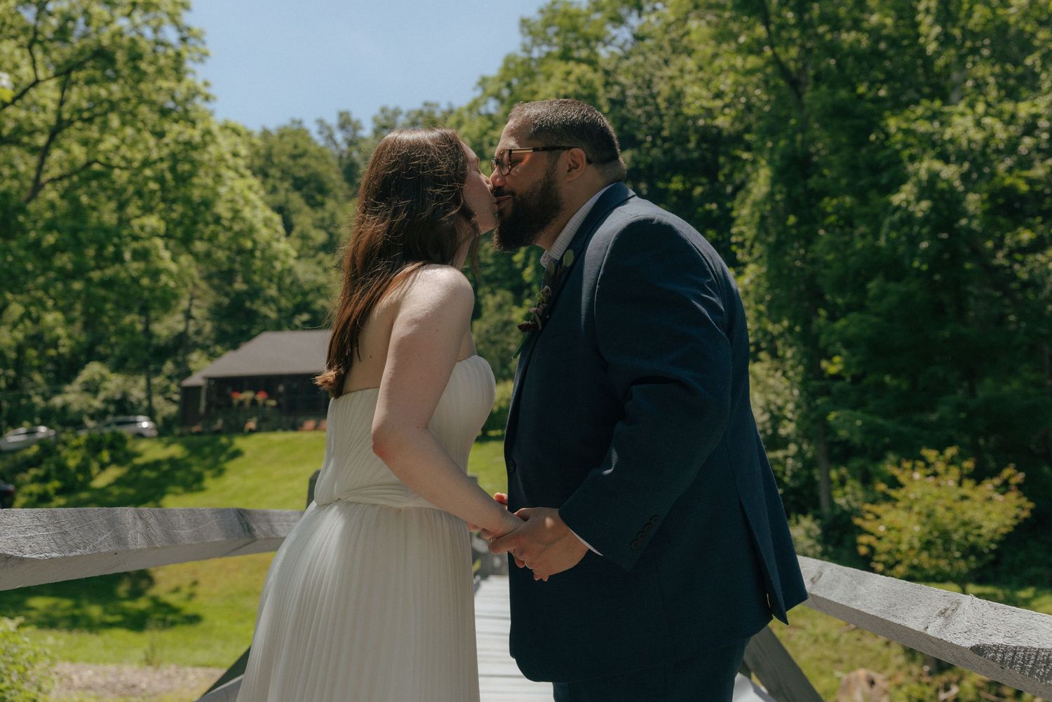 Couple sharing a kiss on a wooden bridge surrounded by lush green trees on a sunny wedding day.