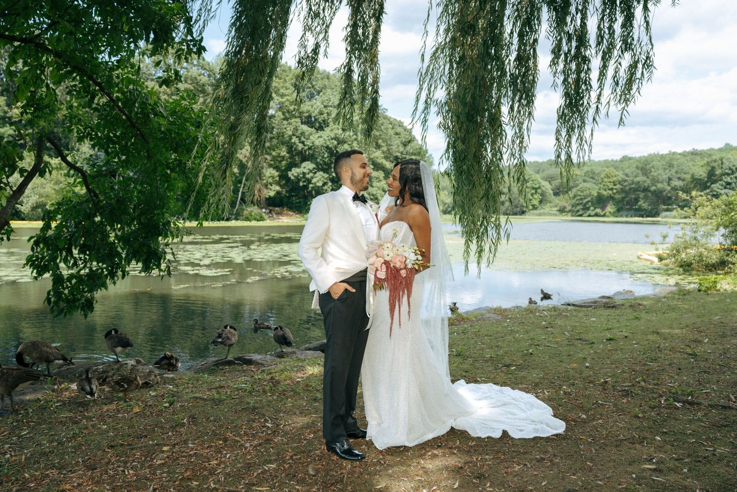 Wedding couple posing by a lake under willow tree with geese in background.