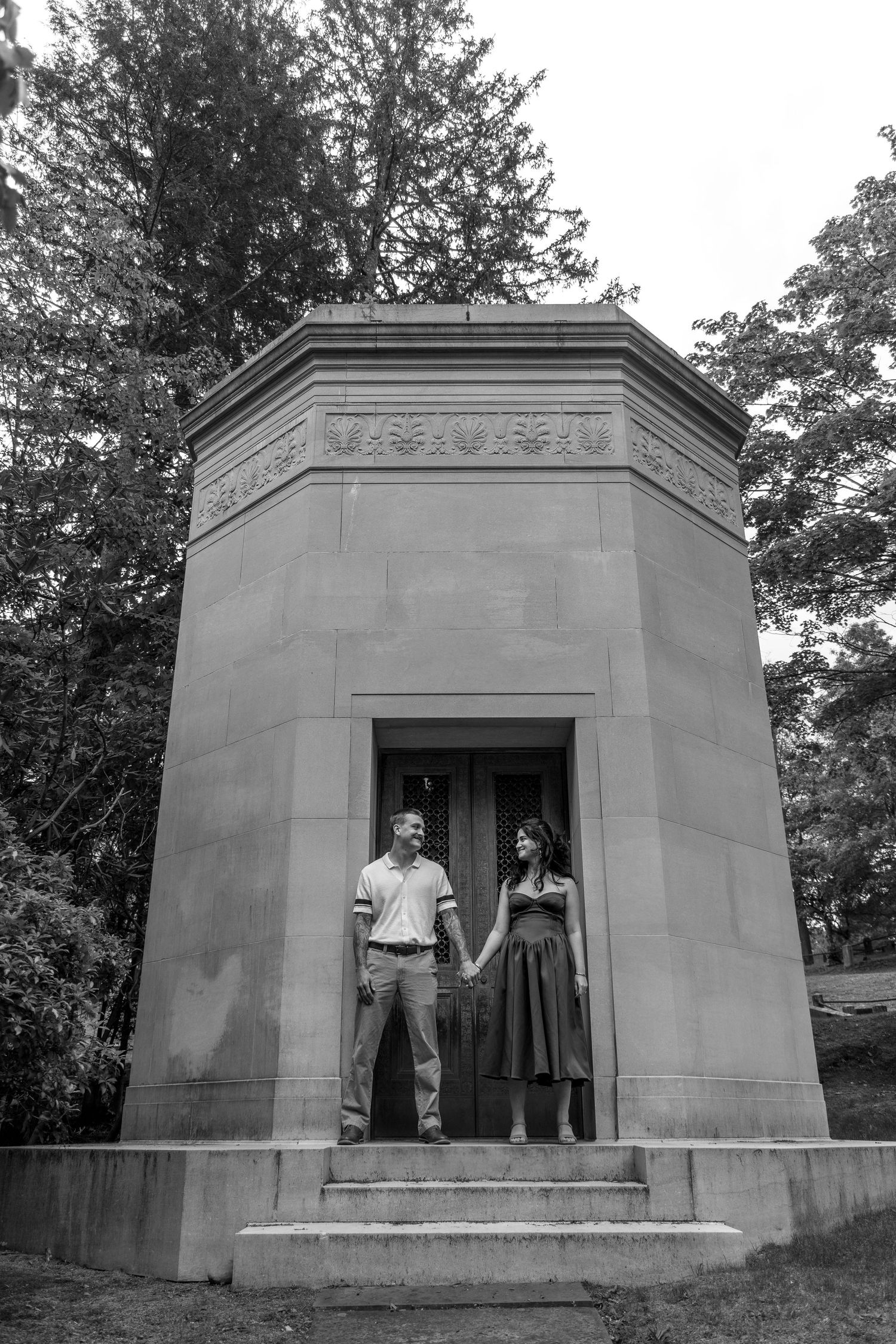 Black and white photo of a couple holding hands on steps of a stone mausoleum surrounded by trees.