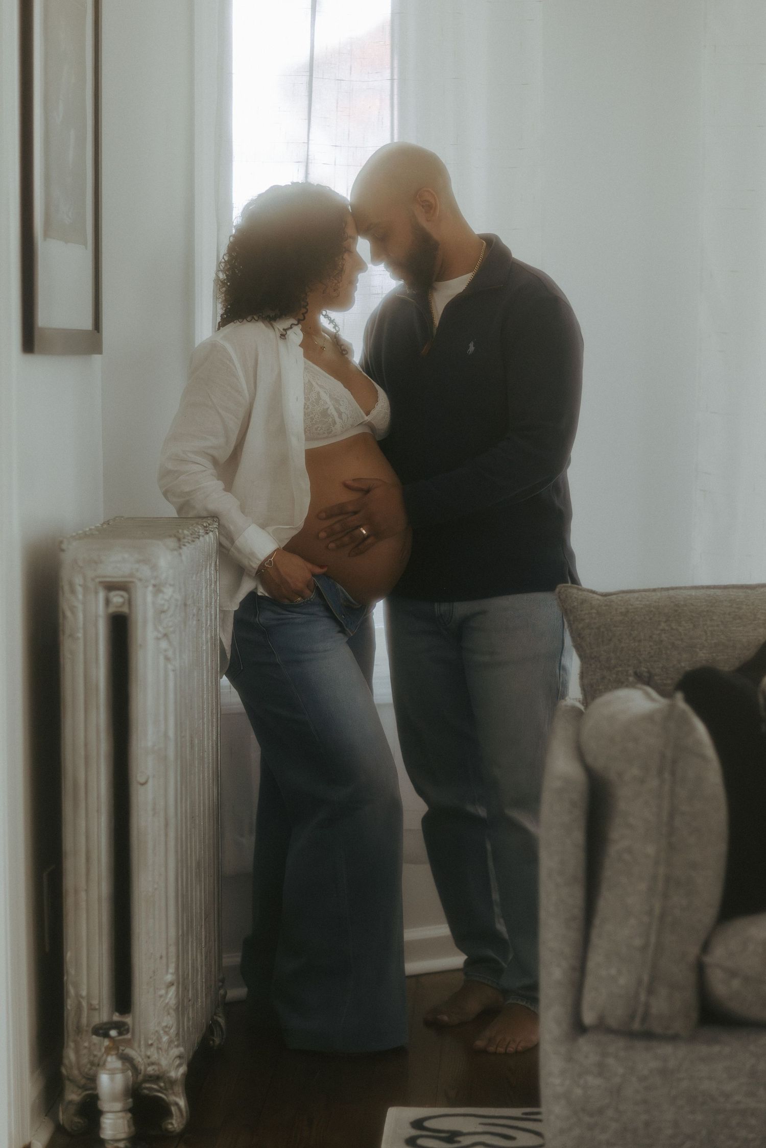 Expecting couple sharing intimate moment by window, pregnant woman in white bralette and jeans.