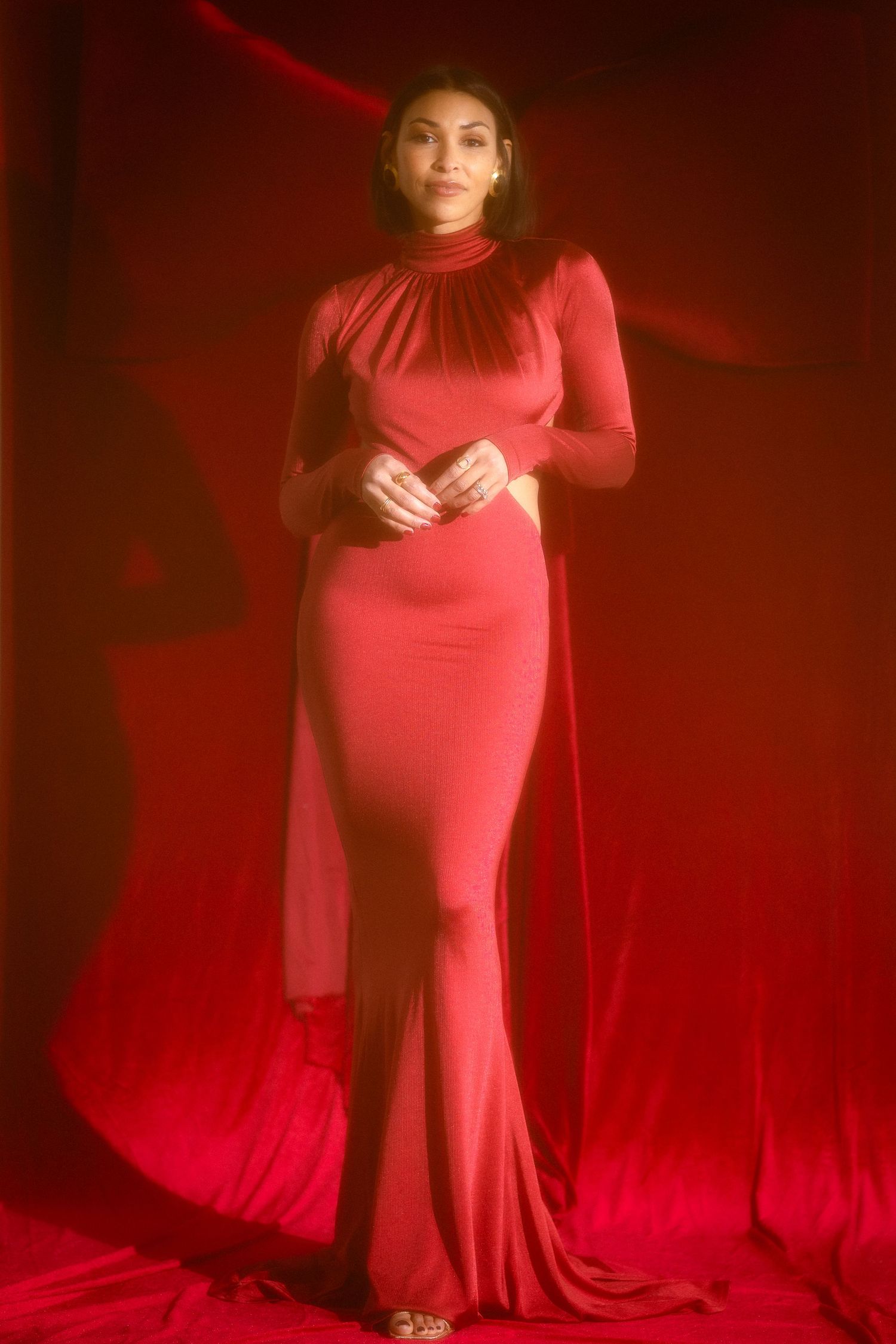 Woman in elegant red long-sleeve backless gown with flowing cape posing on red backdrop.
