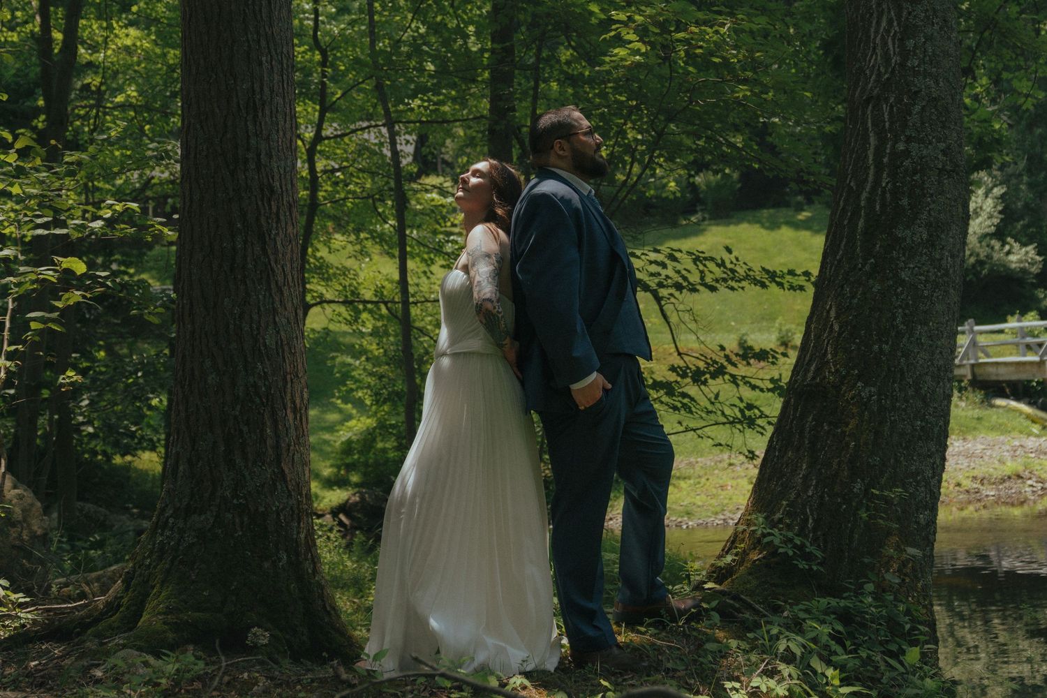 A bride and groom stand back-to-back between trees in a lush green forest during their outdoor wedding.