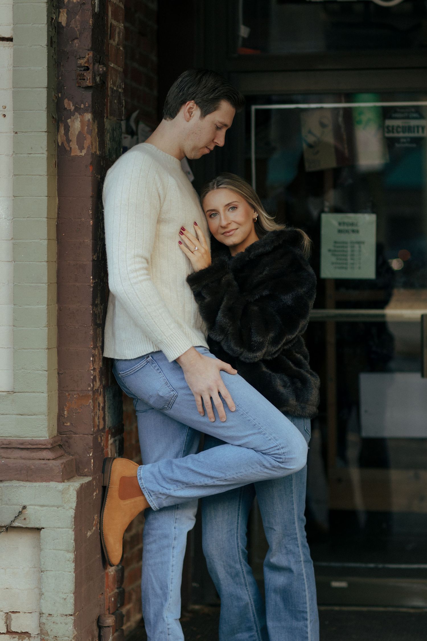 Couple embracing in faux fur coat and cream sweater with jeans, posing against a brick storefront.