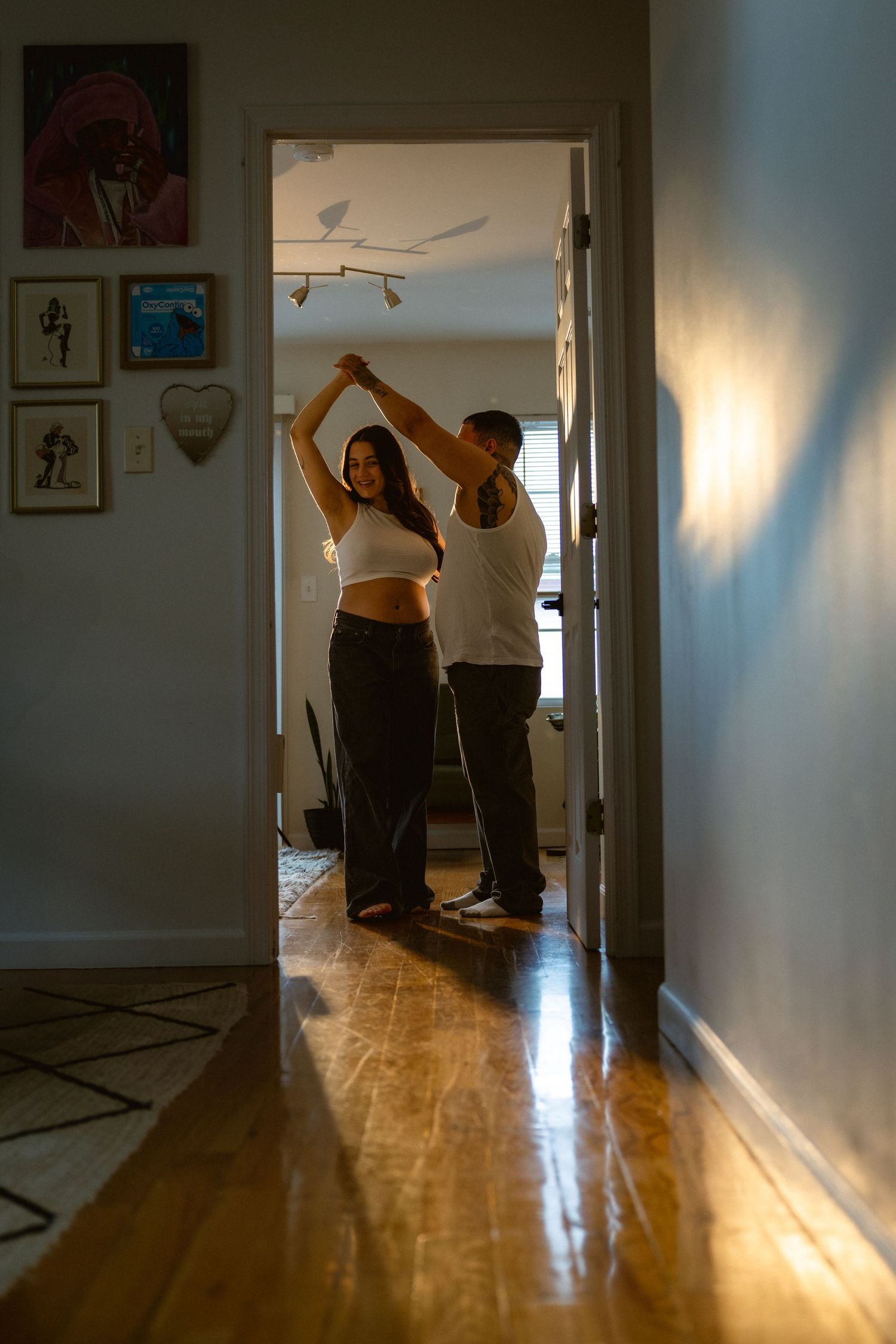 Couple dancing together in a sunlit apartment hallway, with hardwood floors and framed art on the walls.