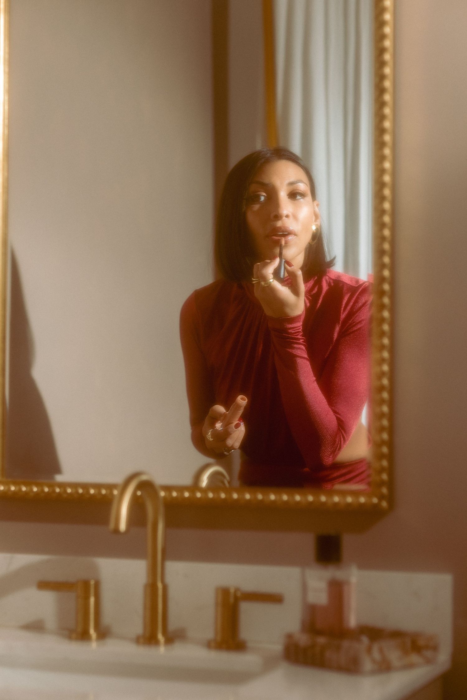Woman in burgundy top applying lipstick captured in ornate gold-framed bathroom mirror.