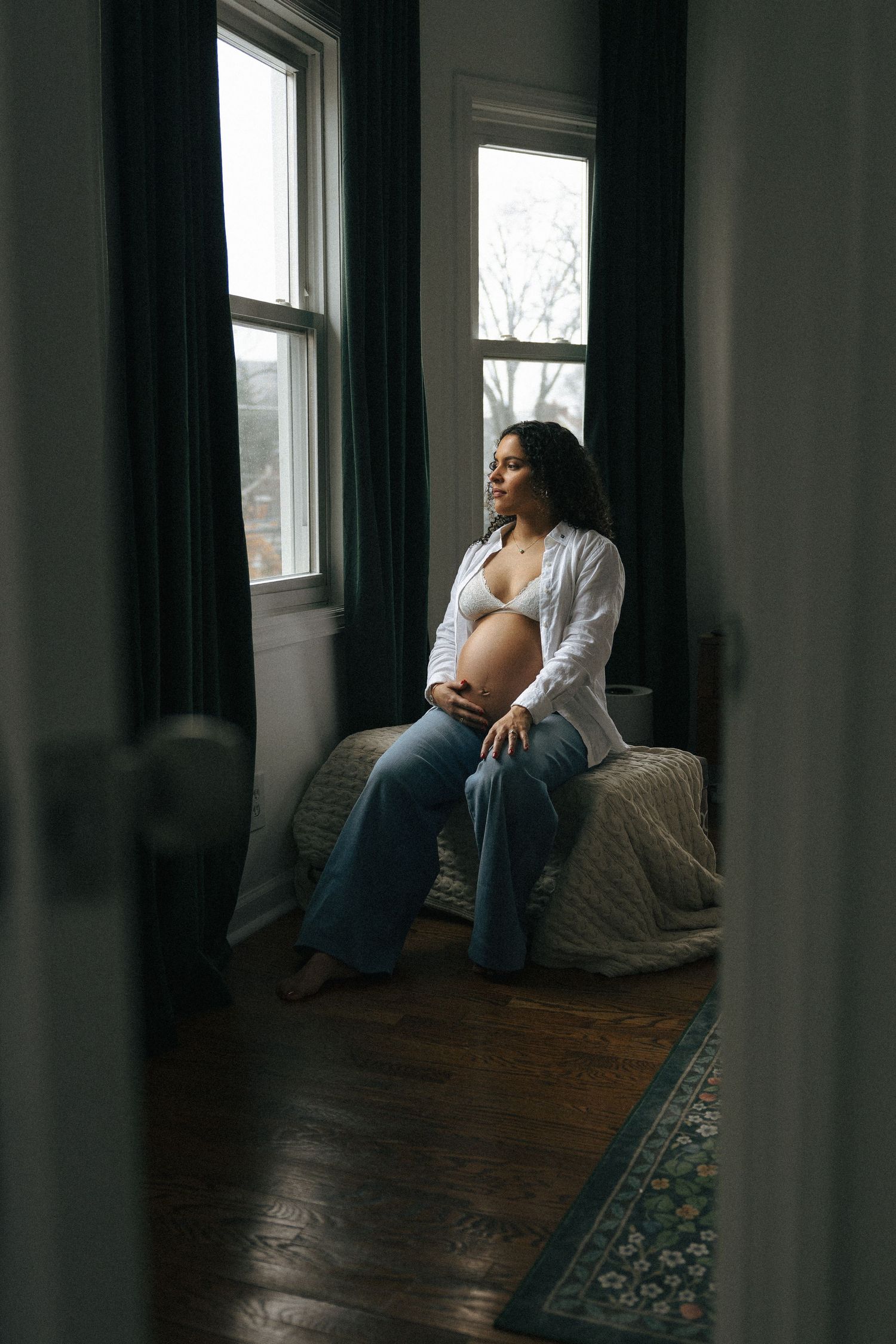Pregnant woman in white shirt and jeans sits on bed by window, cradling baby bump in soft natural light.