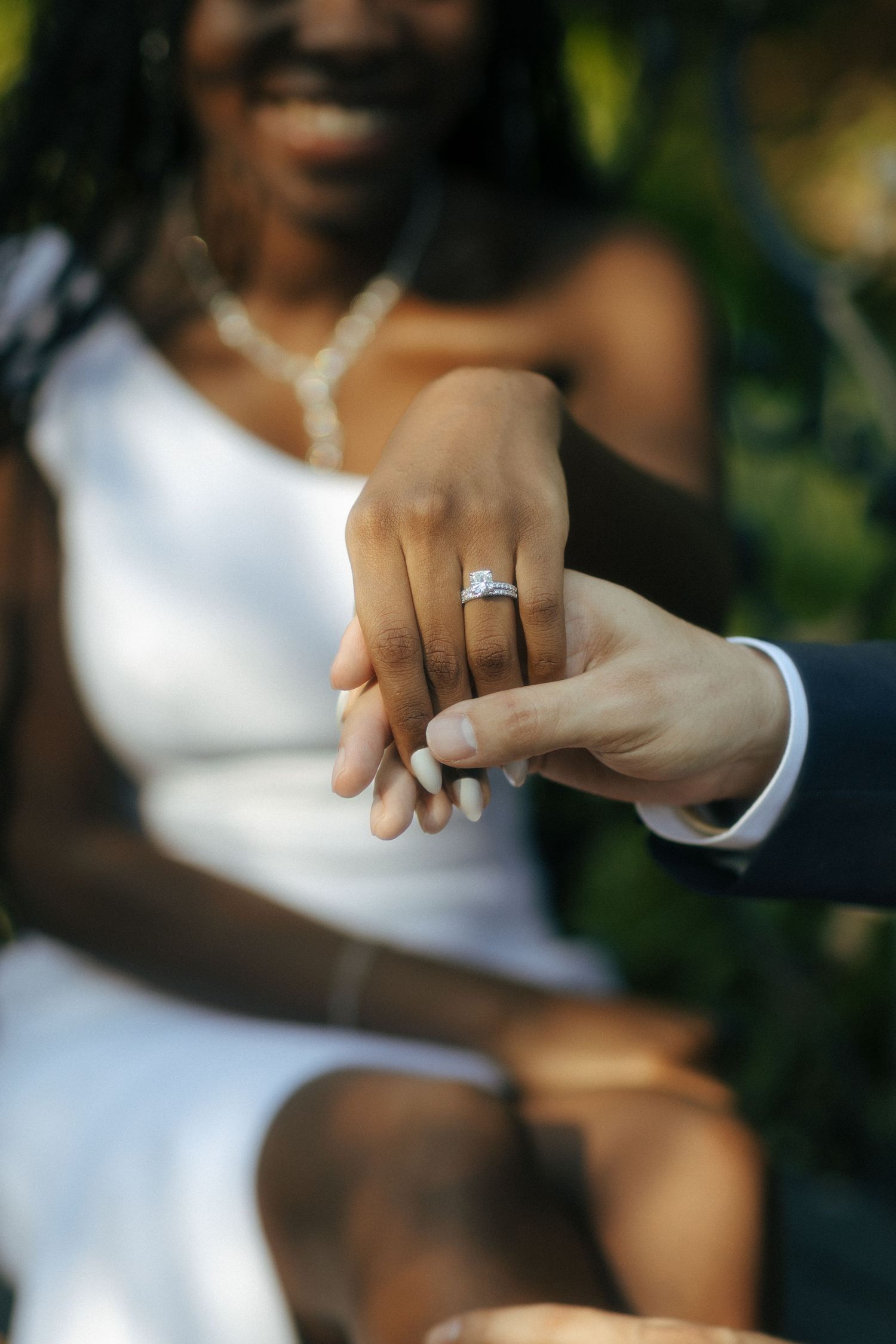 Close-up of bride showing diamond engagement ring while holding groom's hand at outdoor wedding ceremony.