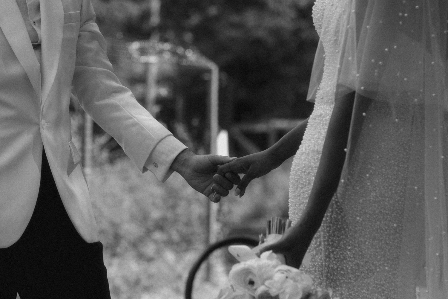 Black and white photo of bride and groom holding hands during wedding ceremony outdoors.