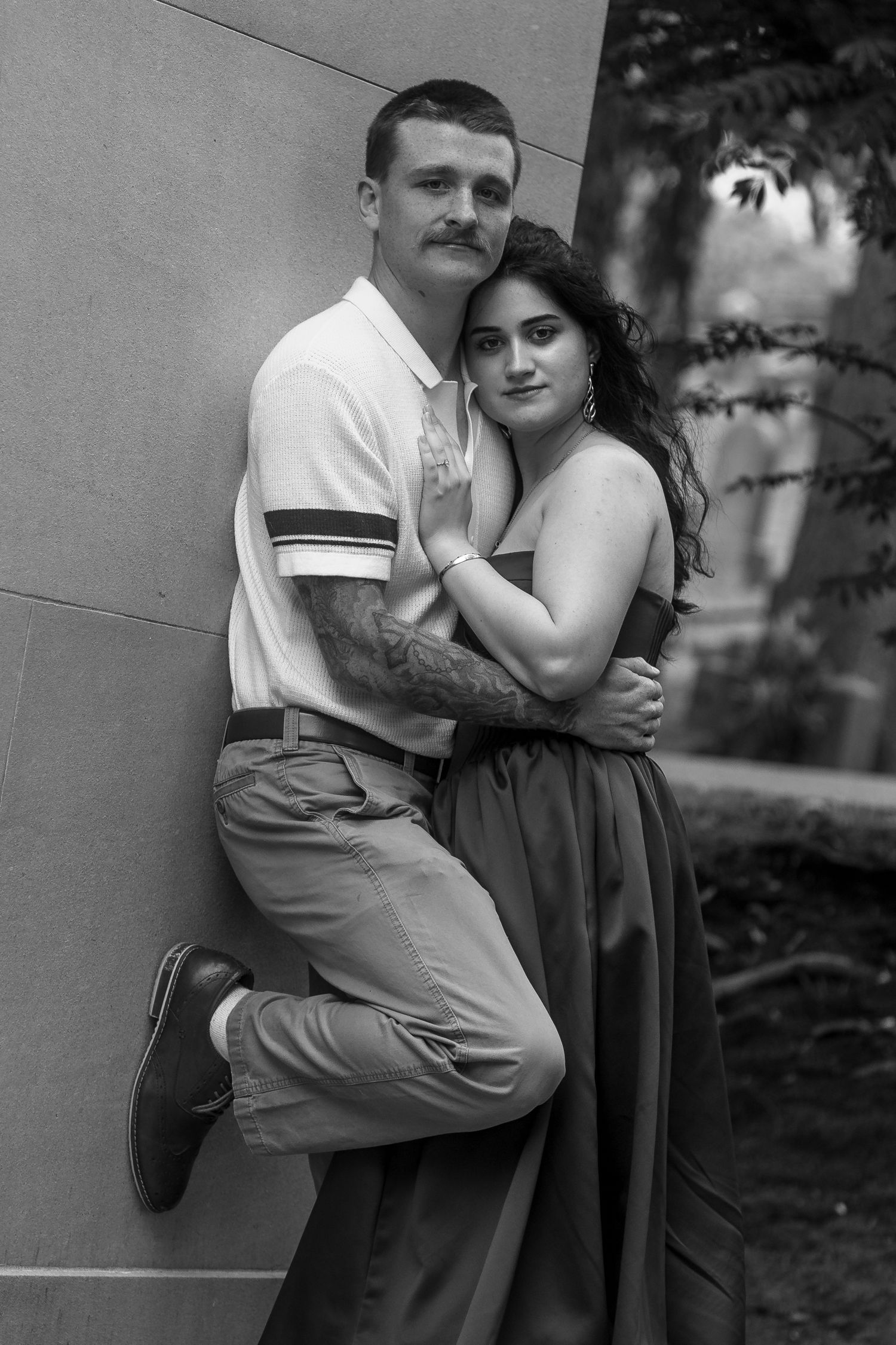 Black and white photo of a couple embracing against a wall, man with mustache and tattoos, woman in strapless dress.