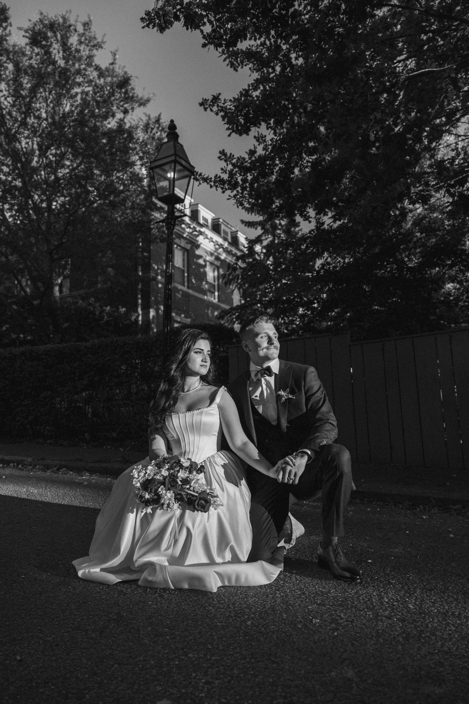 Black and white portrait of bride with bouquet and groom sitting together on street at night.