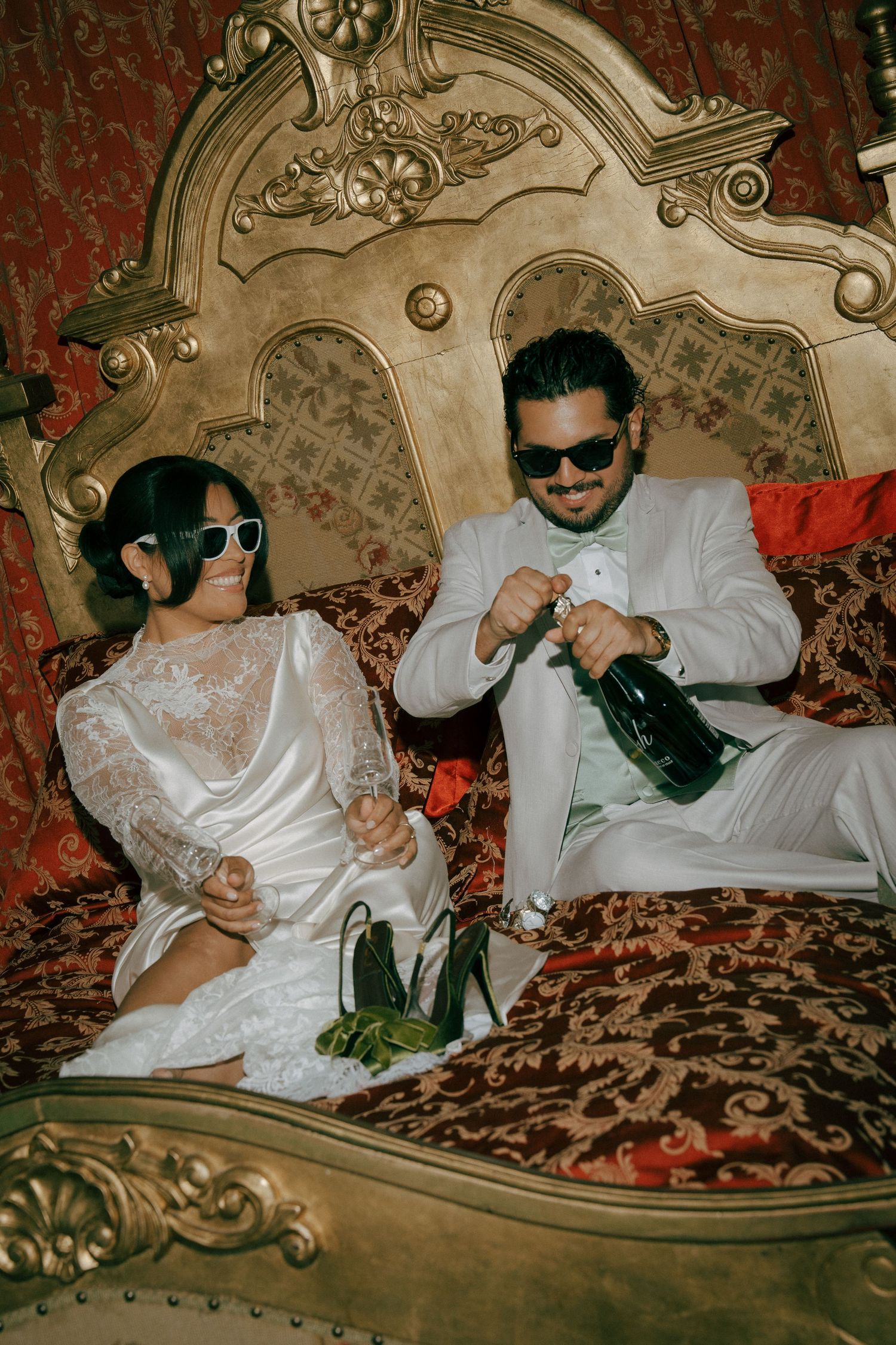 Bride and groom in white outfits and sunglasses opening champagne on ornate golden bed with red velvet decor.