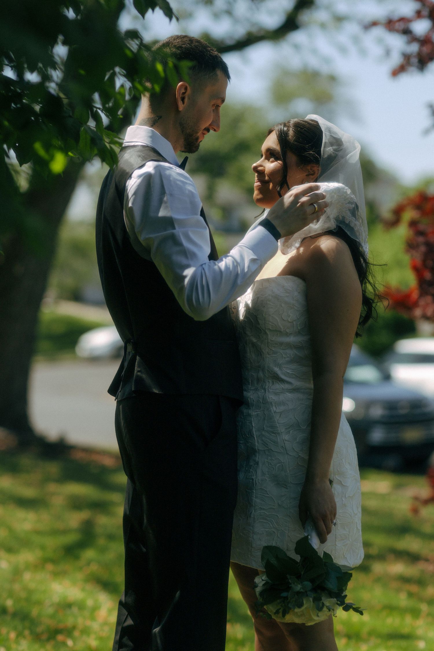 A bride and groom share a tender moment outdoors, with the groom gently holding the bride's face as she smiles up at him.