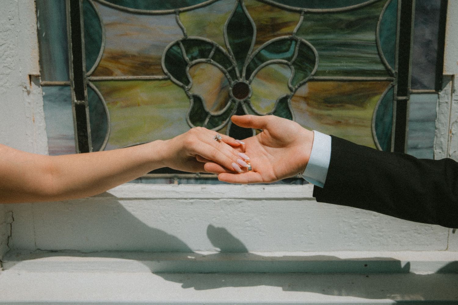 A groom in a suit places a wedding ring on a bride's finger in front of a stained glass window.
