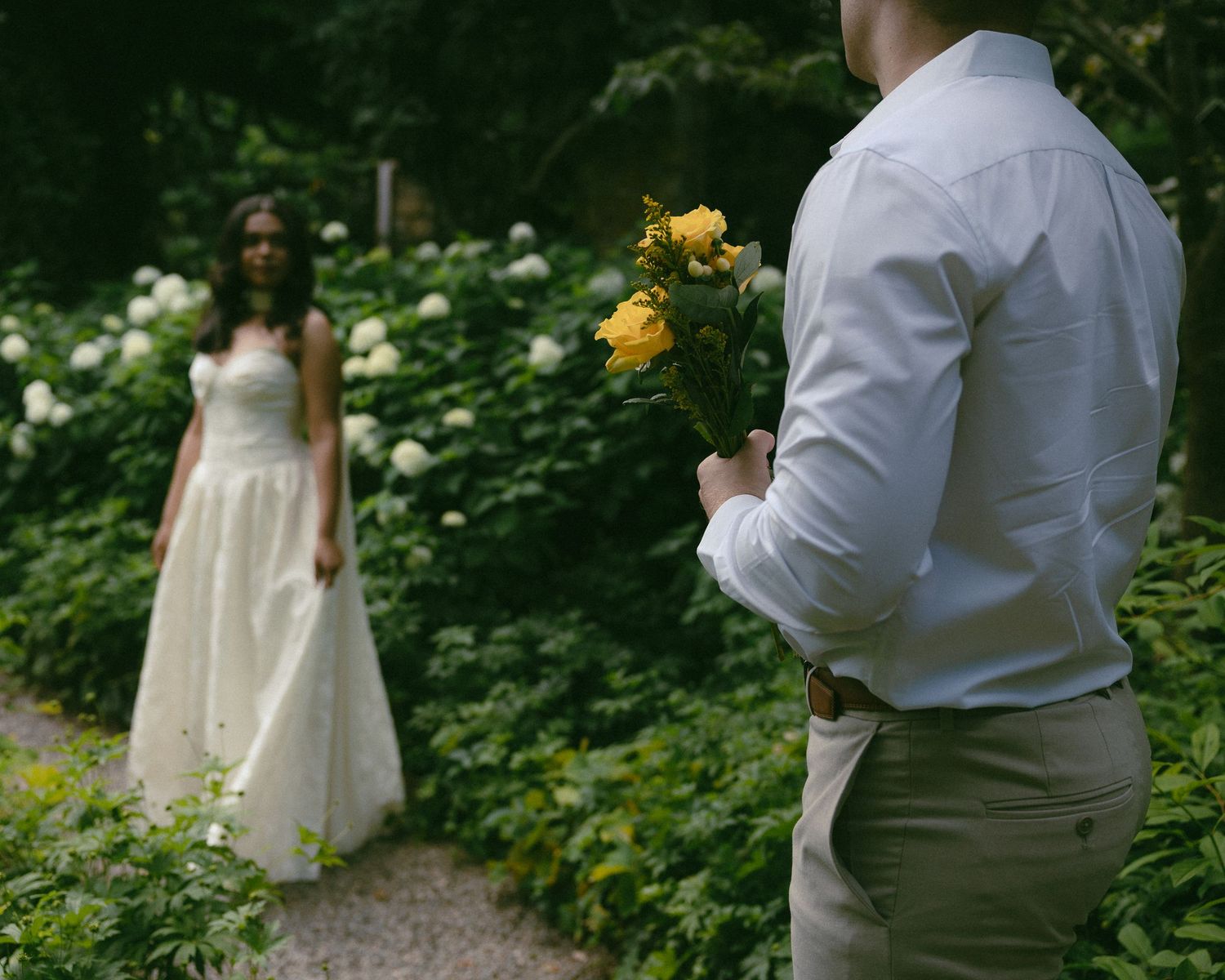 Groom holding yellow roses behind his back surprises bride in white wedding dress on a garden path.