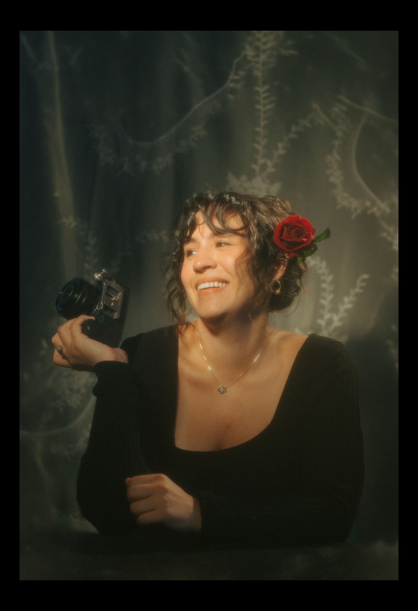 Smiling woman with curly hair and red rose, holding a vintage camera, wearing black top and necklace against dark floral backdrop.
