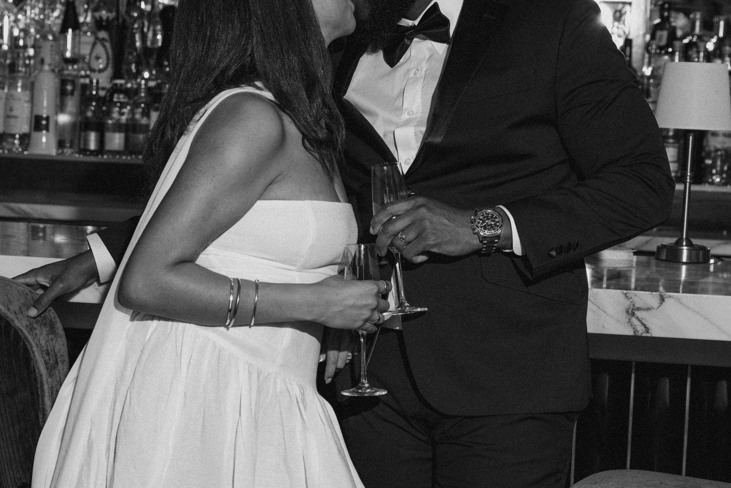 Elegant couple in black and white photo sharing champagne toast, woman in white dress, man in tuxedo at upscale bar.