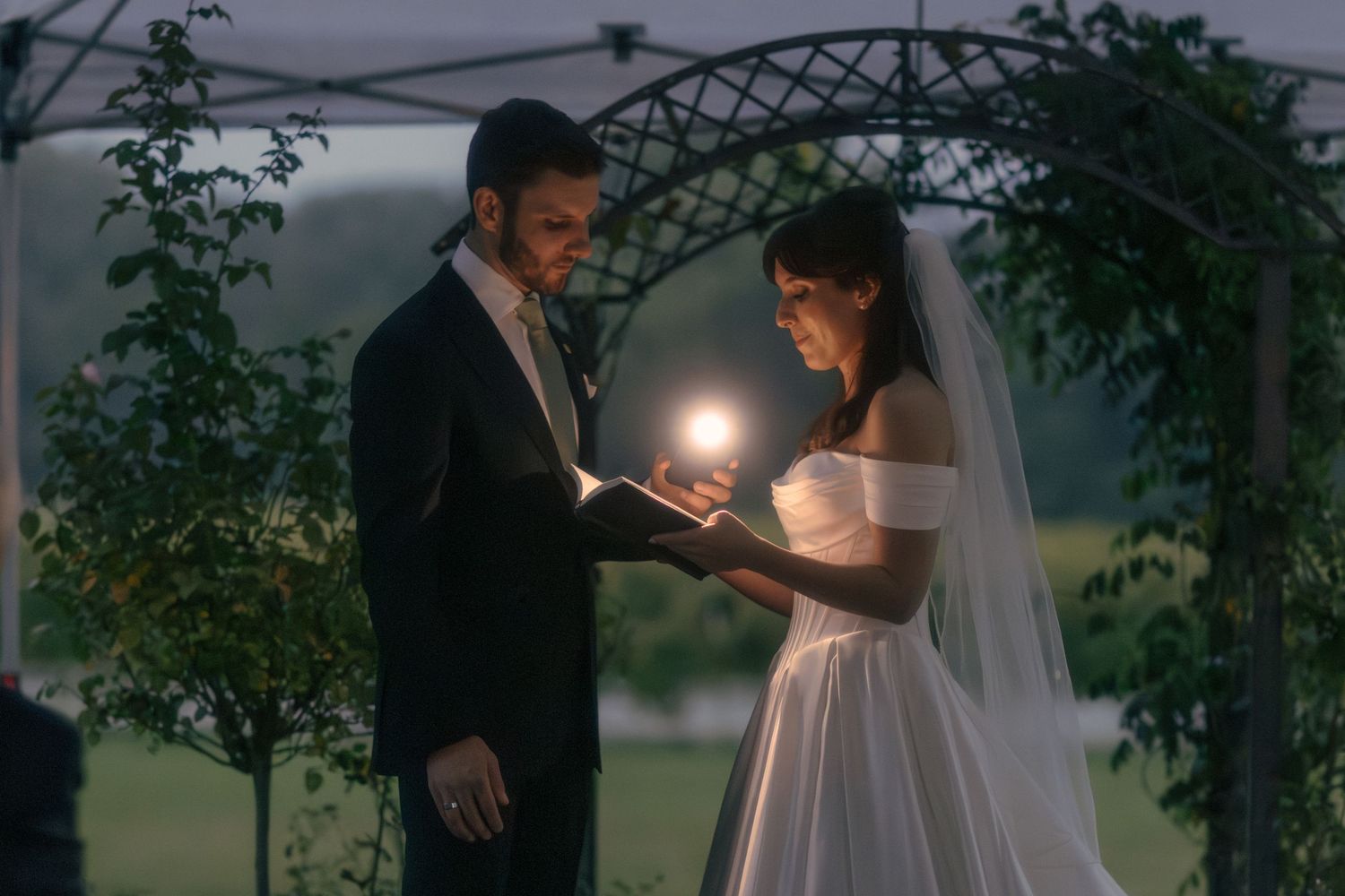 Bride and groom exchange vows by candlelight under an outdoor arch at a romantic twilight ceremony.
