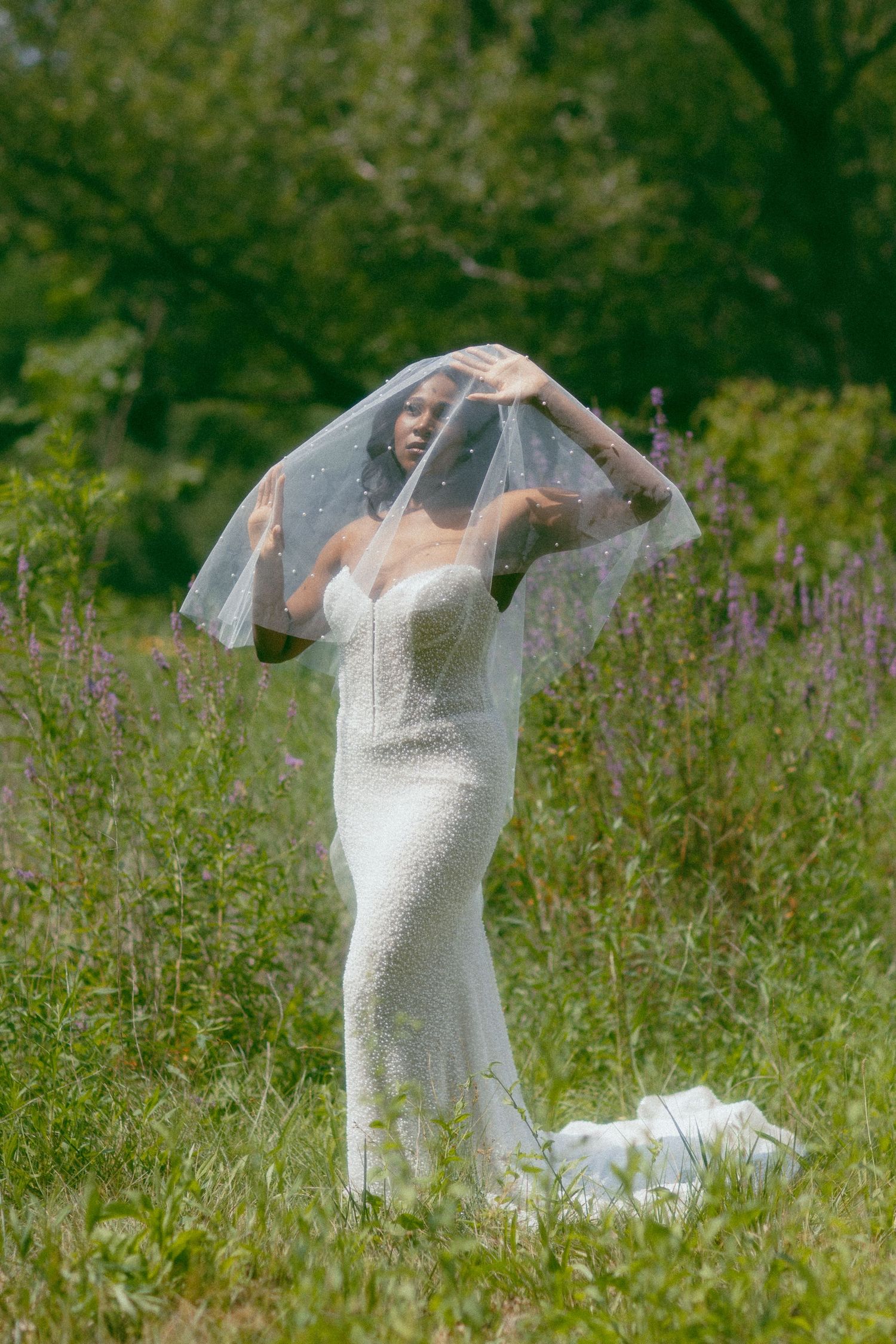 Black bride in fitted white lace wedding dress lifts veil outdoors in lush green meadow.