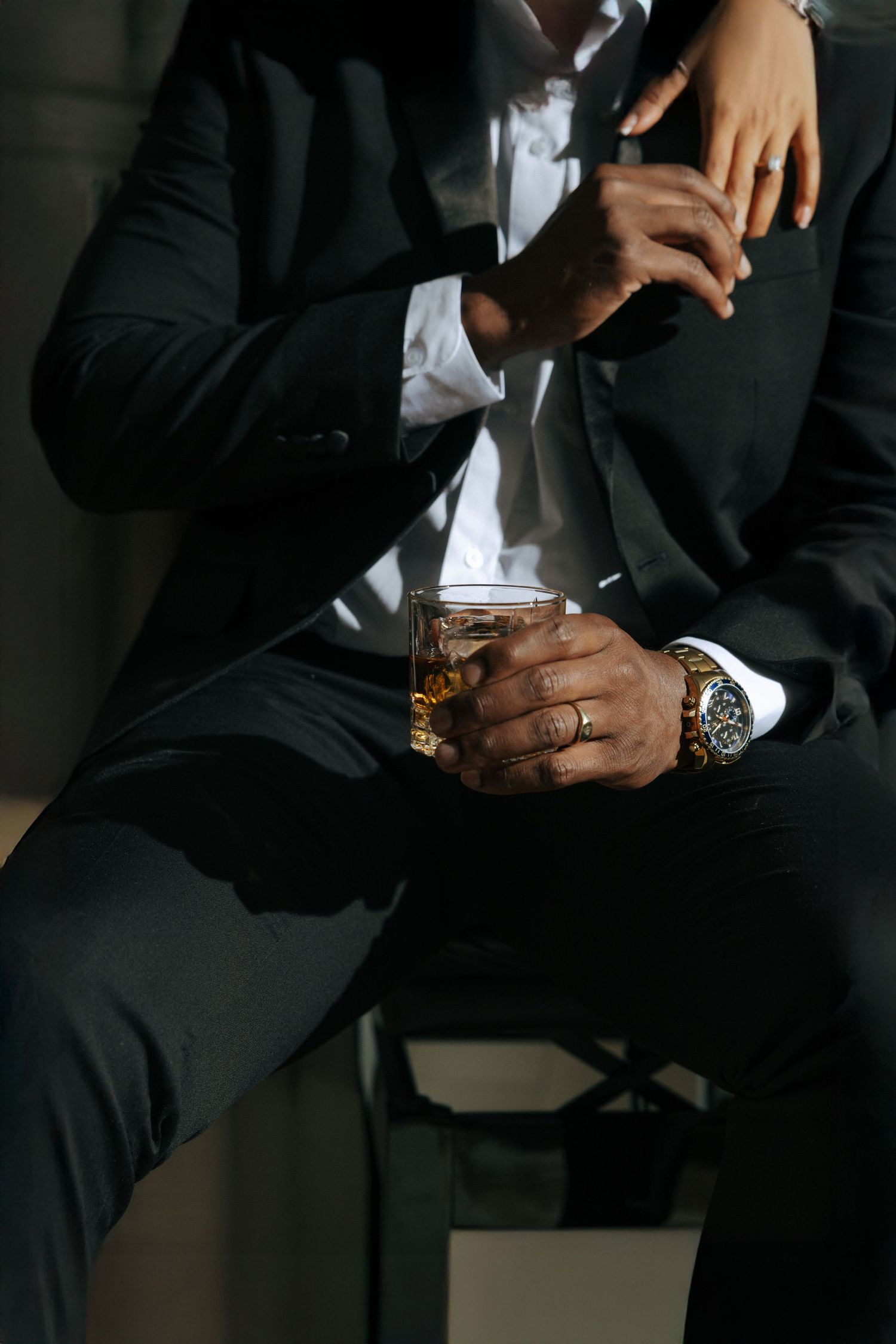 Man in black suit holding whiskey glass with gold watch, ring visible, woman's hand on shoulder in moody lighting.