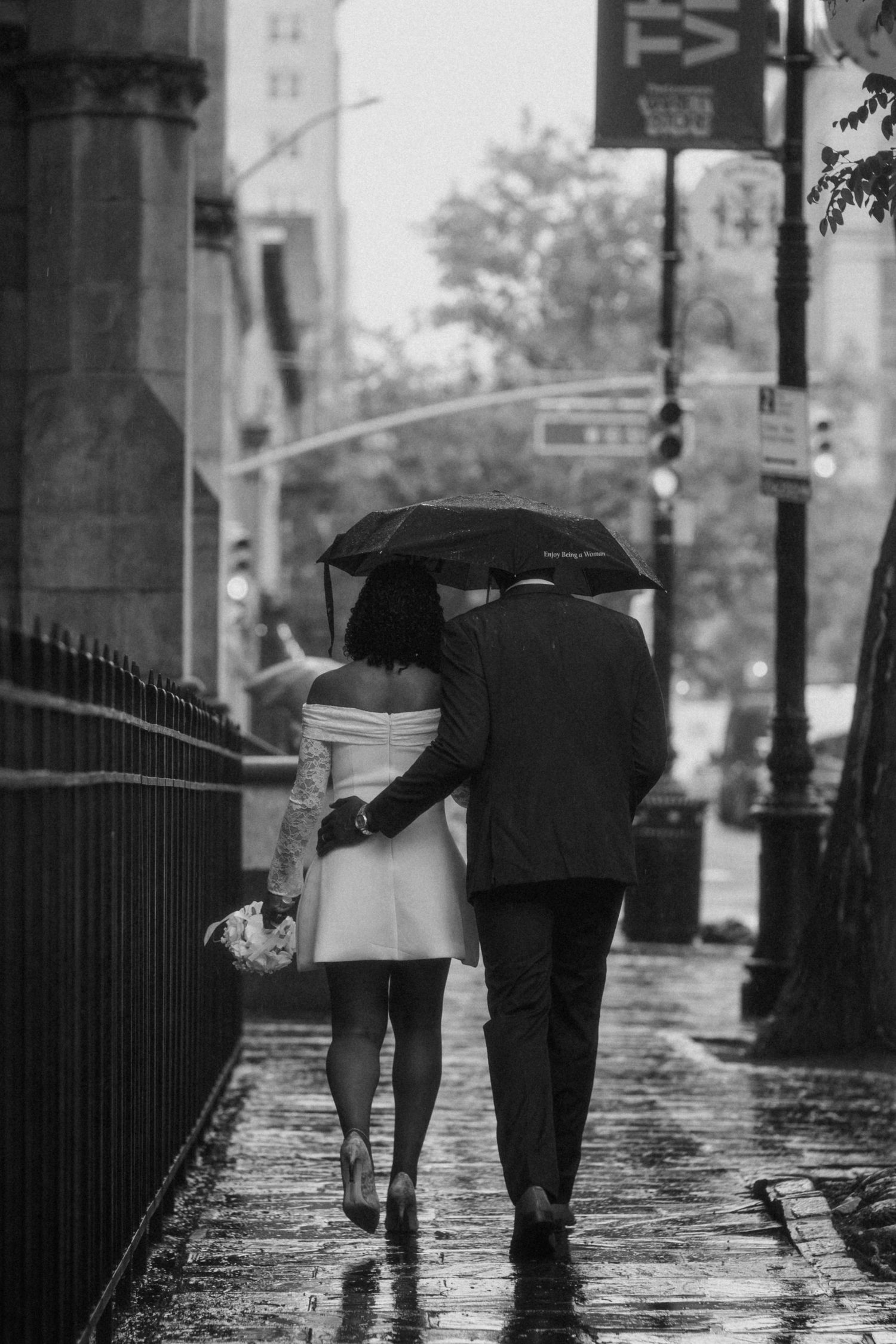 Black and white photo of a newlywed couple walking under an umbrella in the rain on a city sidewalk.