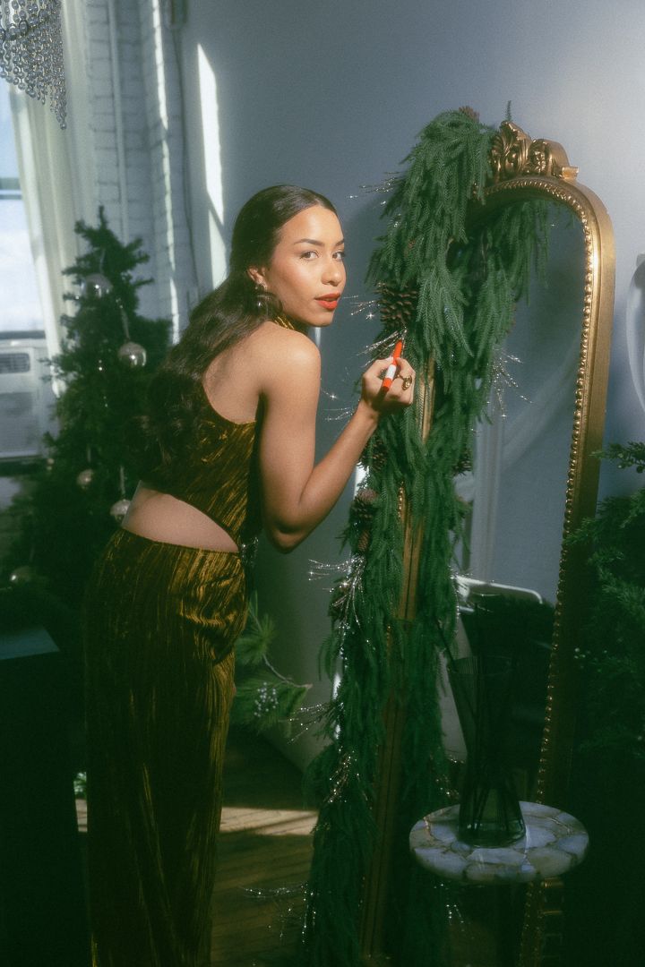 Woman in gold velvet dress decorating with greenery near ornate mirror in holiday styled room.