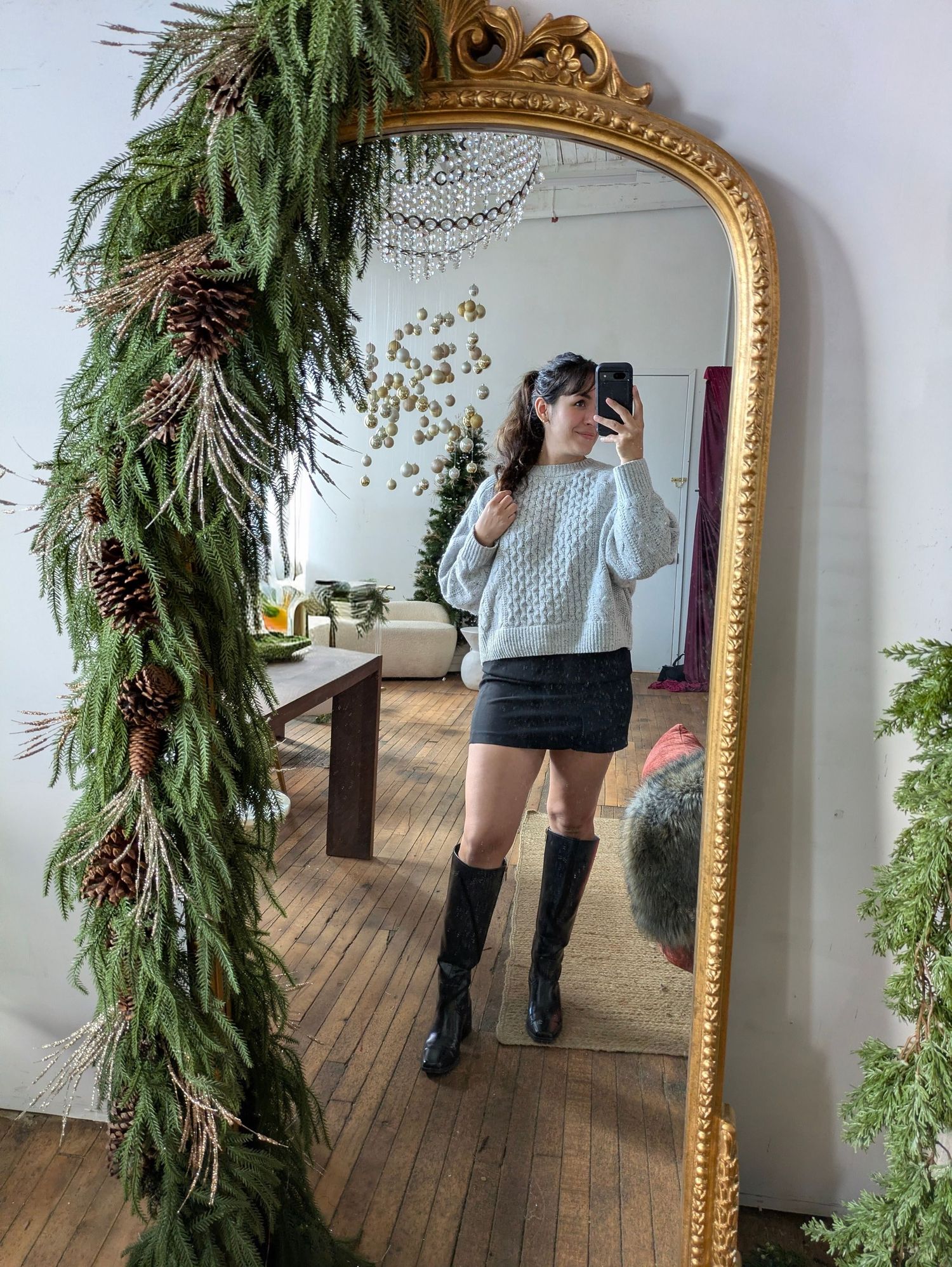 Woman taking a mirror selfie in a light blue sweater and black mini skirt with tall boots.