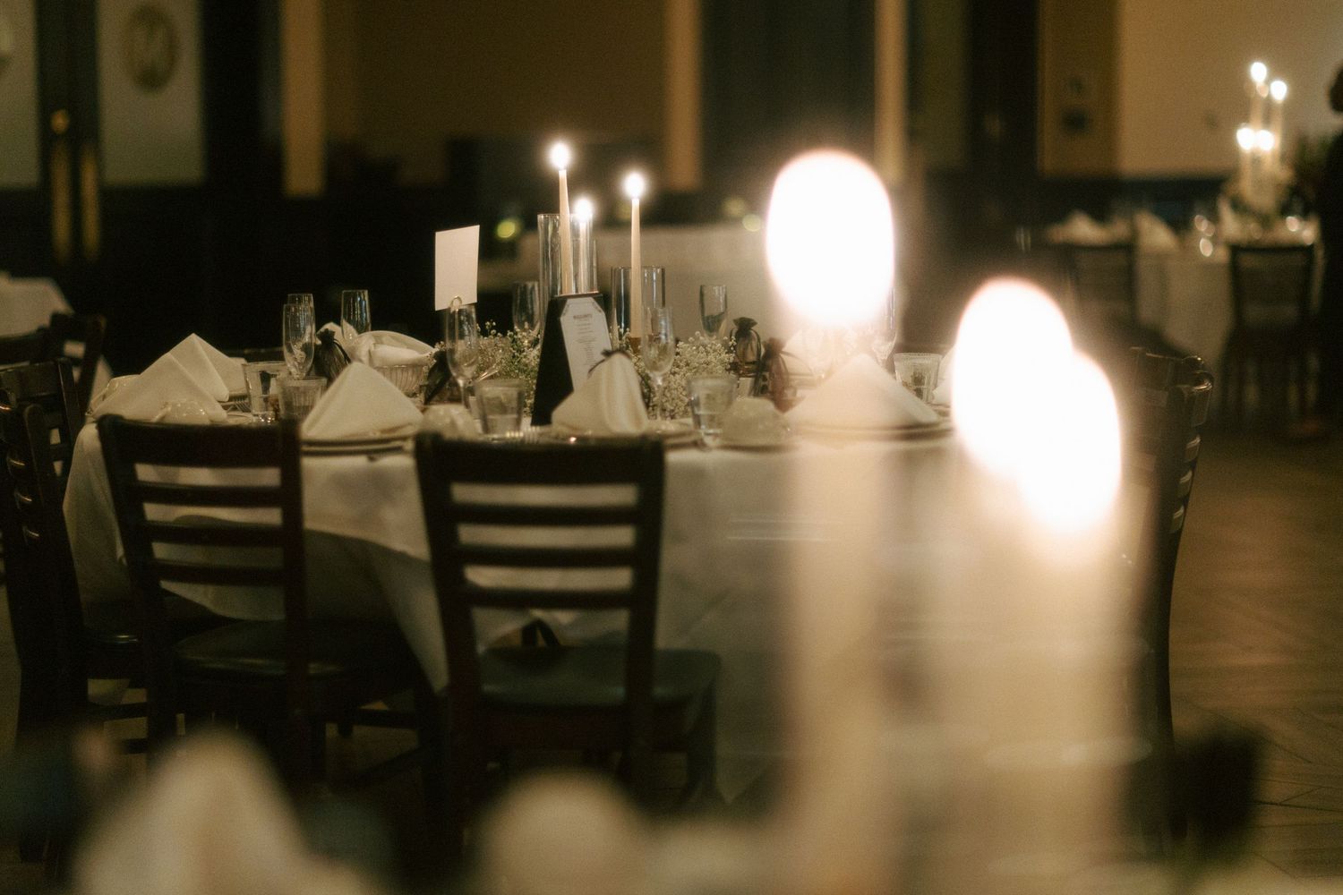 Romantic banquet table setting with white linens, folded napkins, and glowing candles in a dimly lit reception hall.