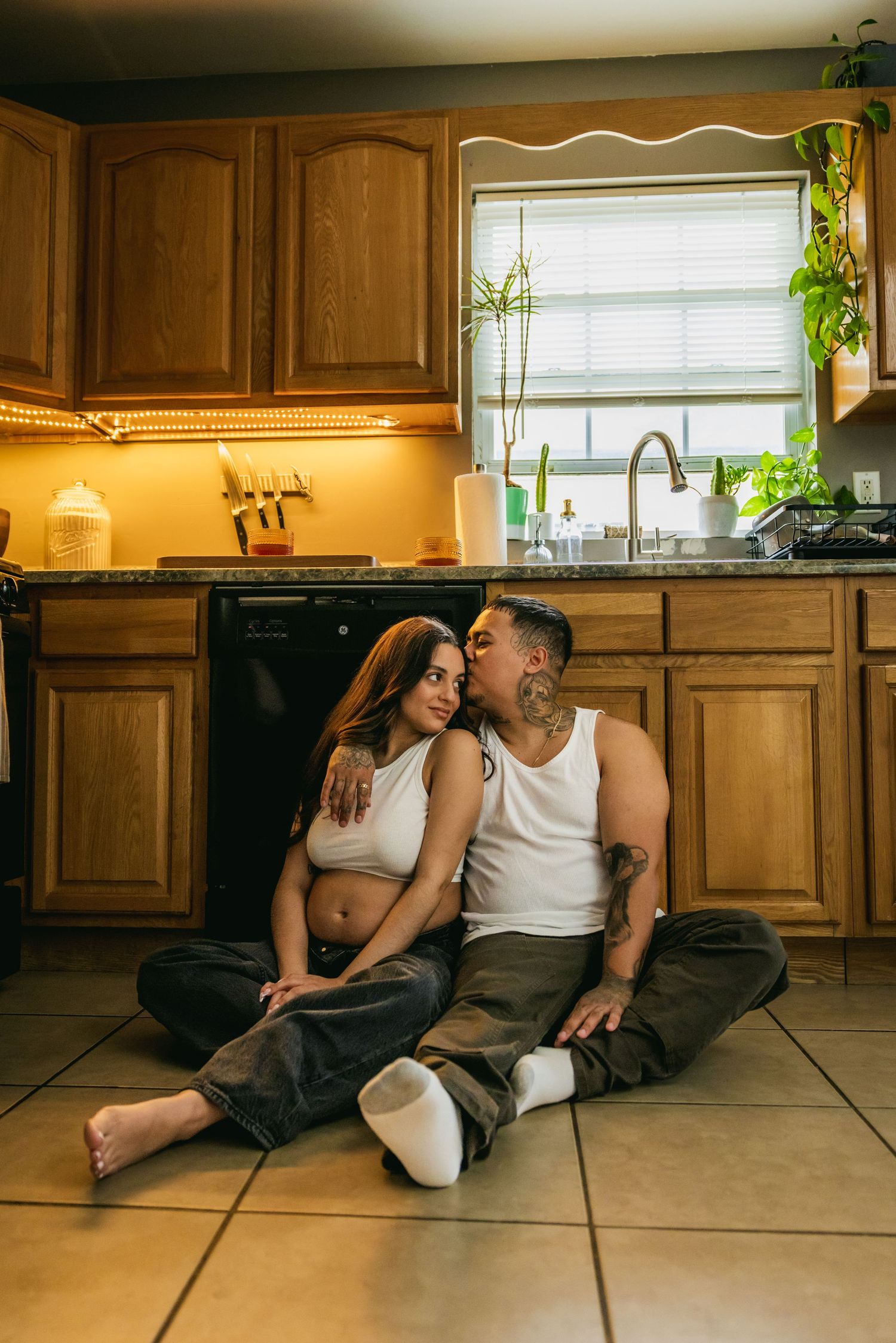 A couple sits on a kitchen floor, man kissing pregnant woman's cheek, both in casual white tops.