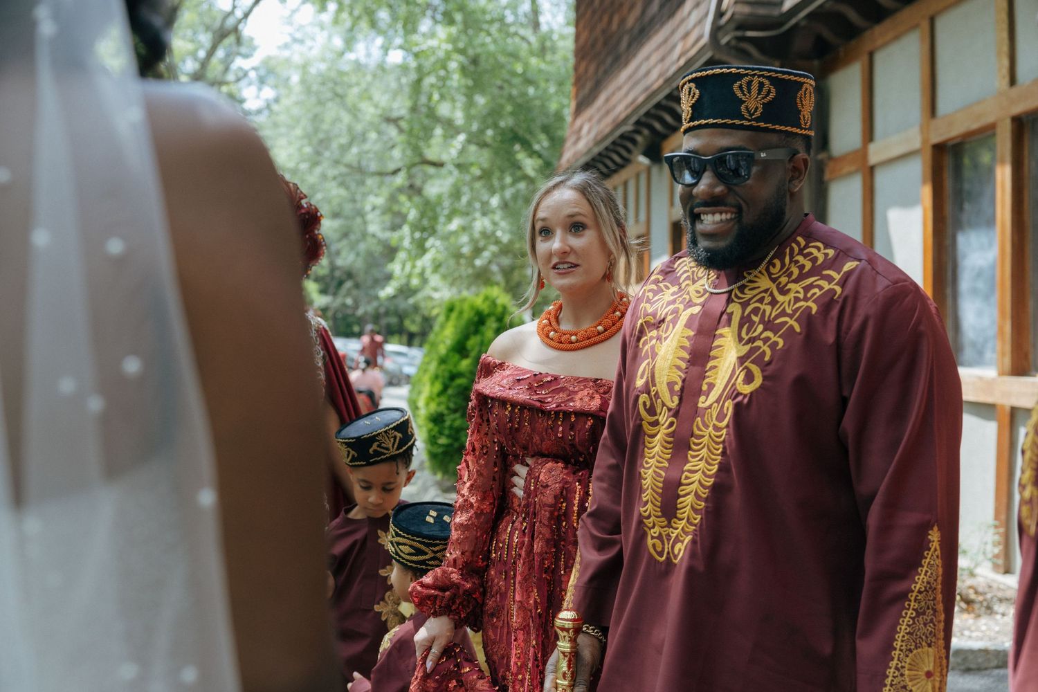 Wedding guests in traditional African attire smiling outside a wooden building.