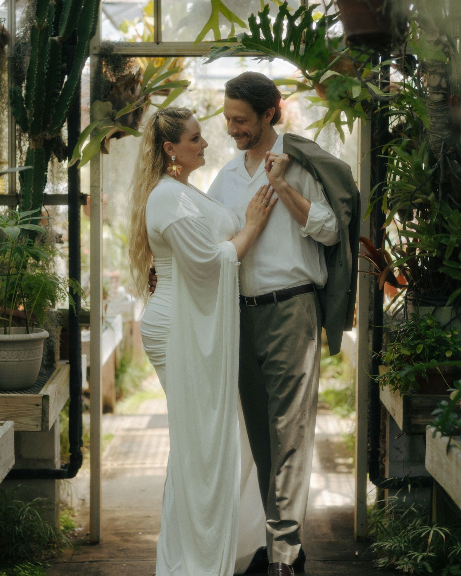 Couple in white dress and gray suit embracing in a lush greenhouse surrounded by tropical plants.