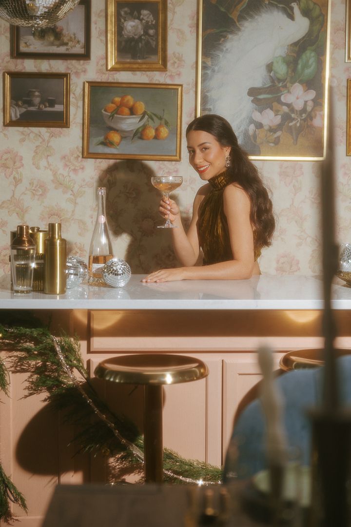 Elegant woman in dark halter top holding champagne glass at bar counter in floral wallpapered room.