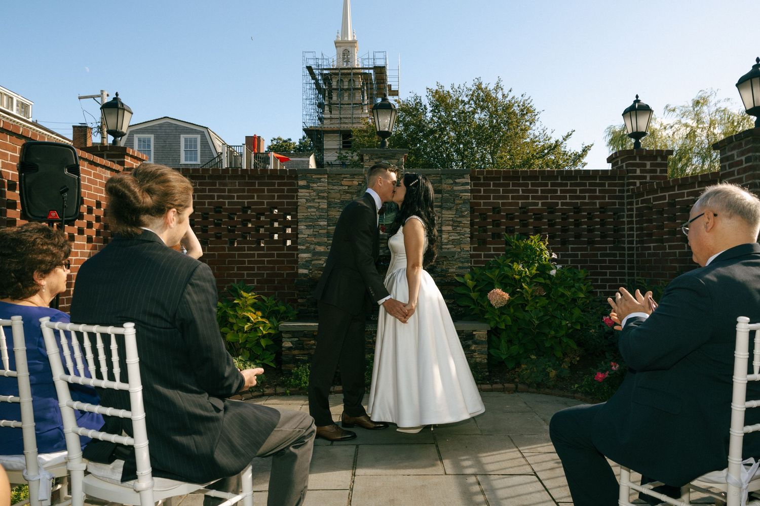 Bride and groom share first kiss at outdoor rooftop wedding ceremony with brick wall backdrop.