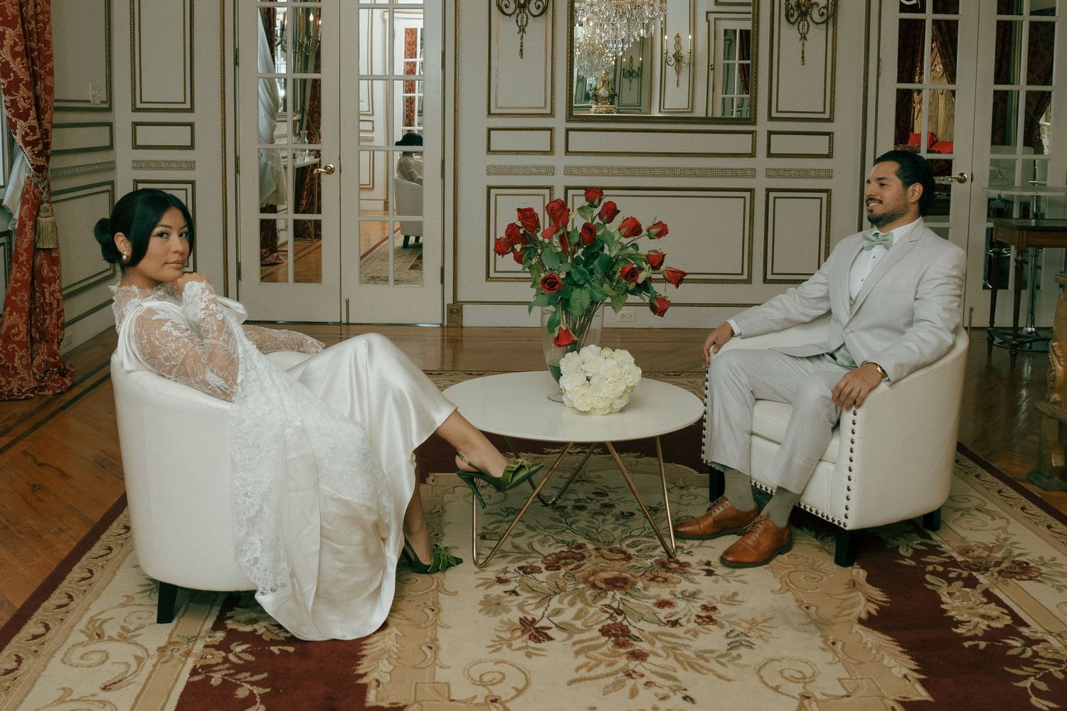 Bride and groom in elegant attire sit in white armchairs with red roses on a coffee table in a luxurious room.