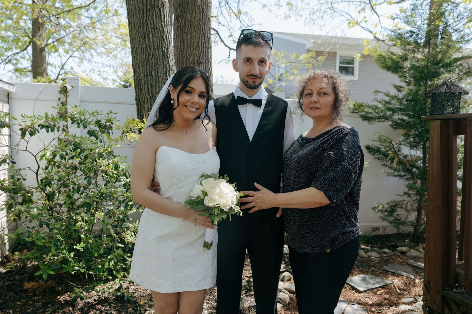 Bride in white strapless dress holding bouquet poses with groom in tuxedo and older woman in backyard garden setting.